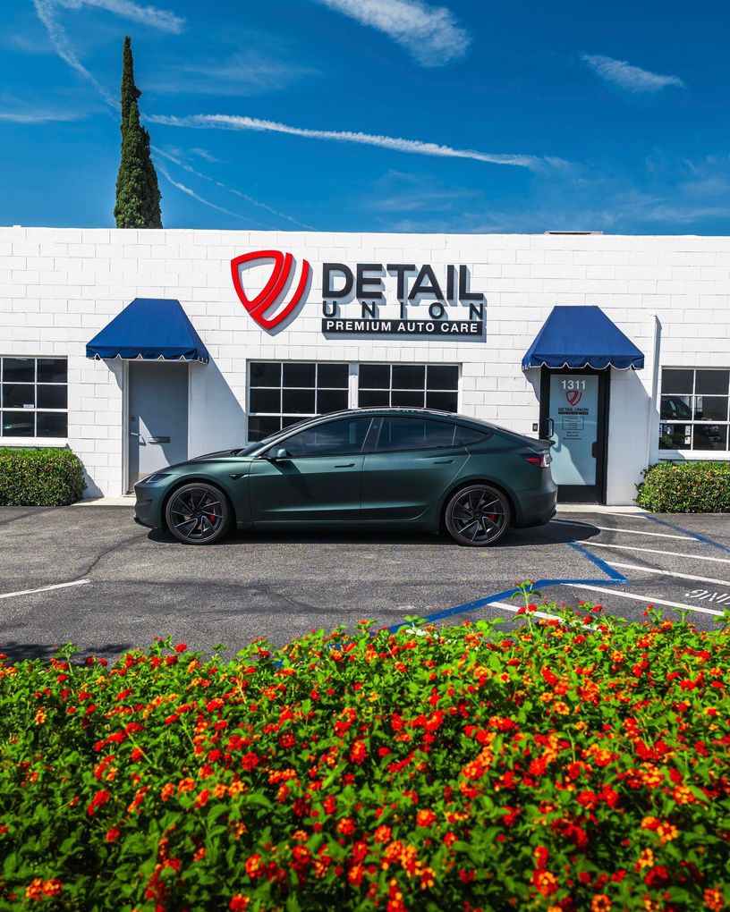 Dark green sedan parked outside a detail shop with blue awnings and bright flowers in front