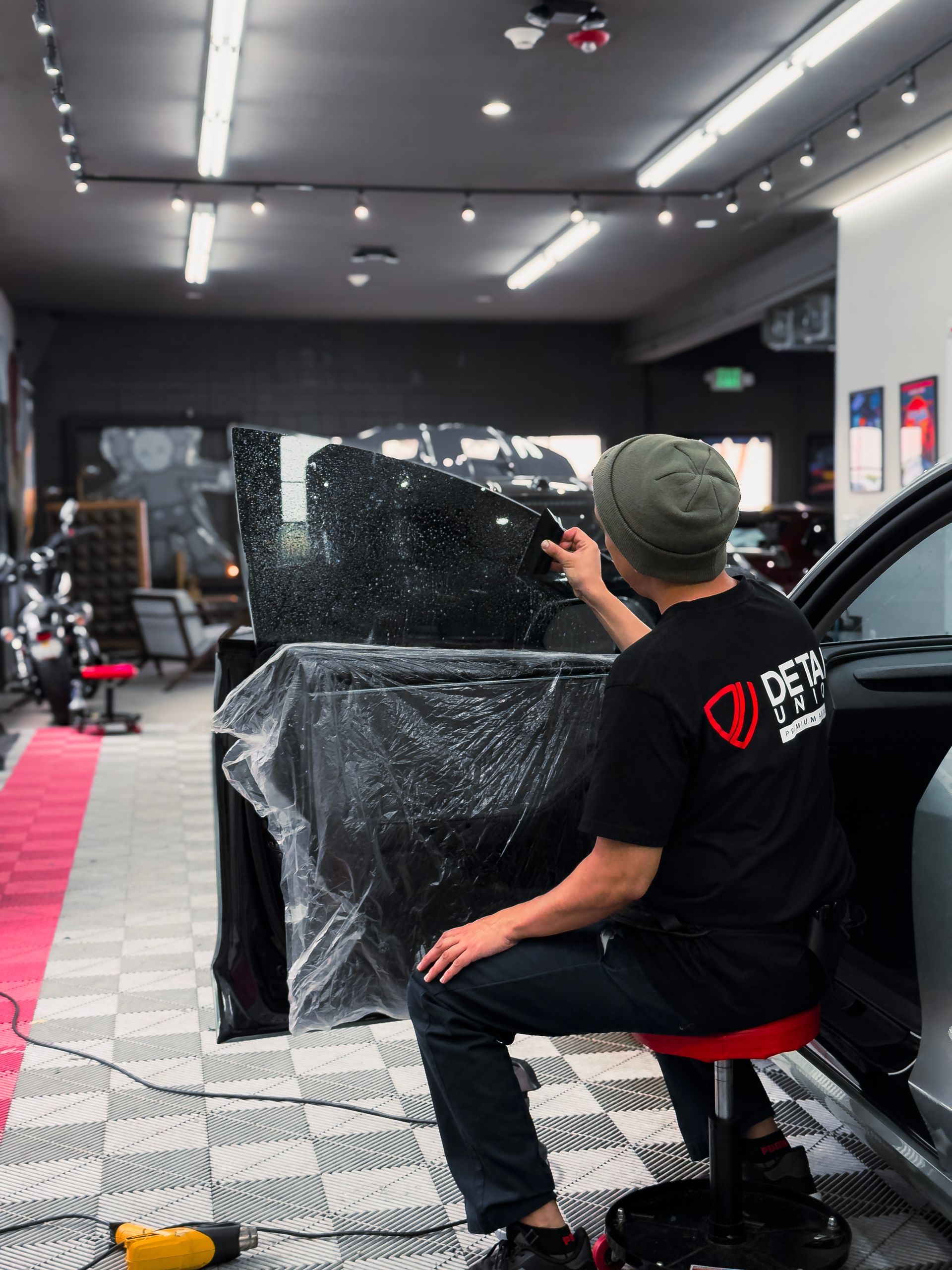 Person spray painting a gold SUV with clear protective plastic on a workshop floor
