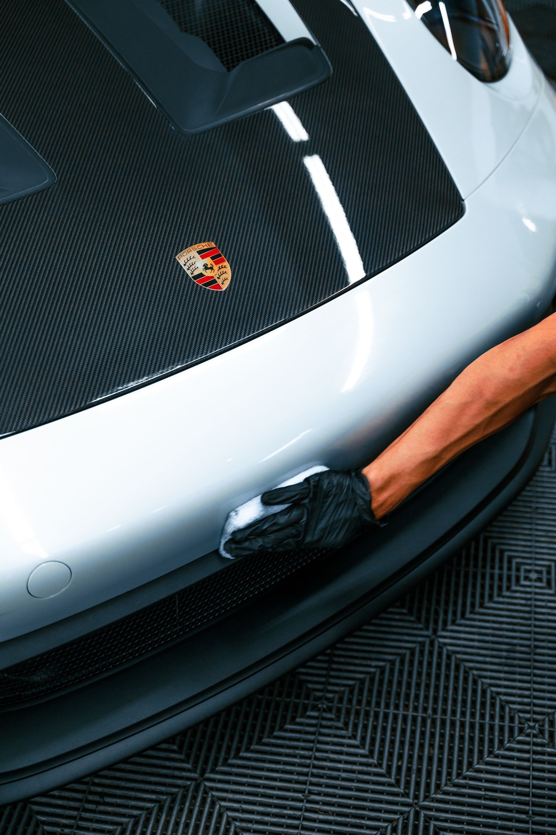 Gloved hand polishing the front bumper of a silver Porsche on a carbon-fiber floor