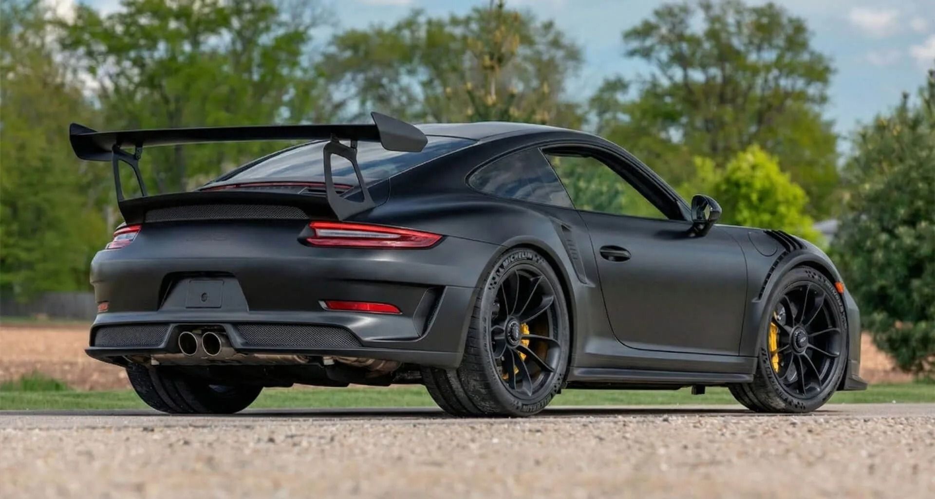 Matte black sports car with a large rear wing parked on a road, side view in a green outdoor setting