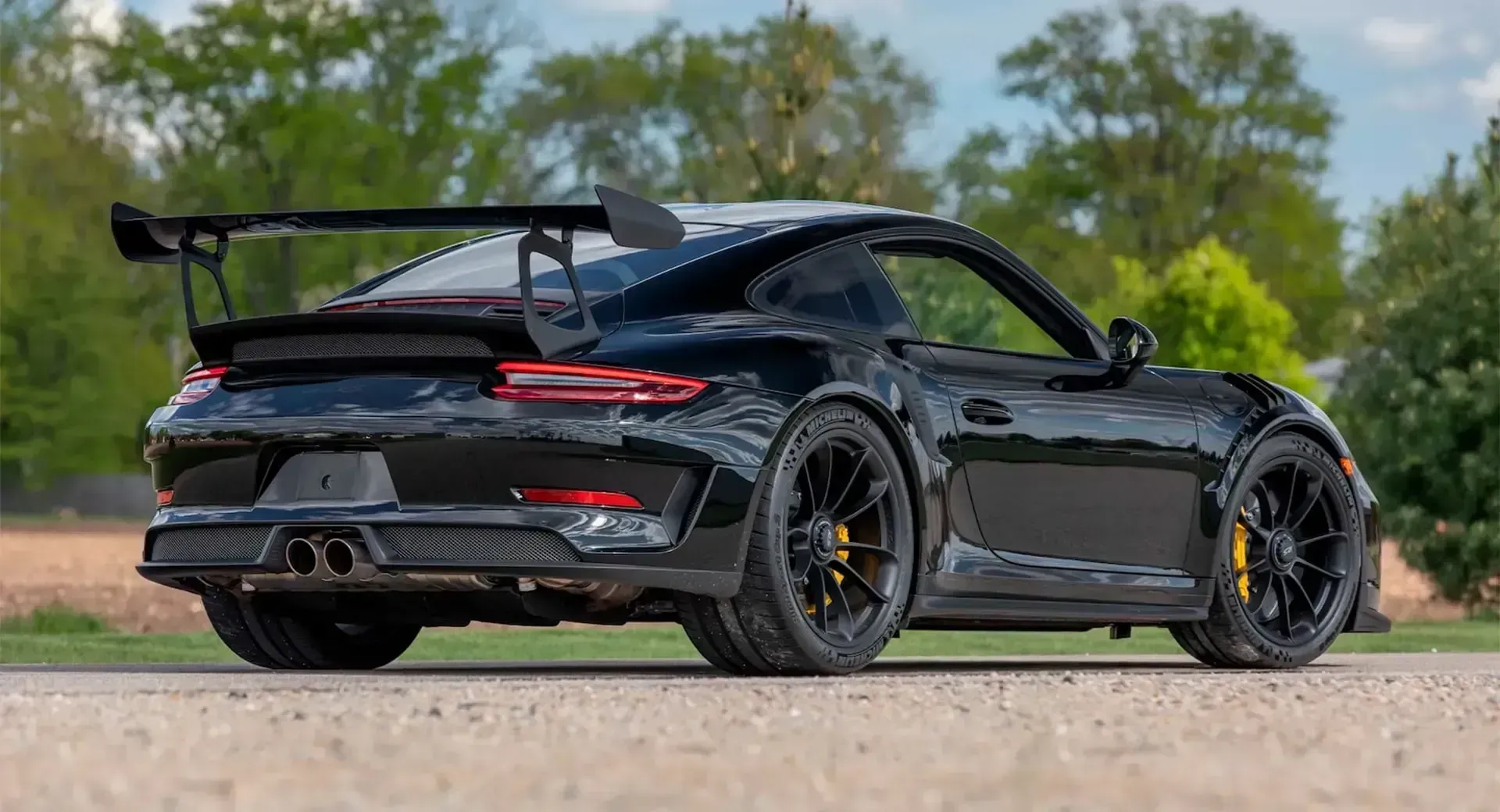 Matte black sports car with a large rear wing parked on a dirt road with trees in the background