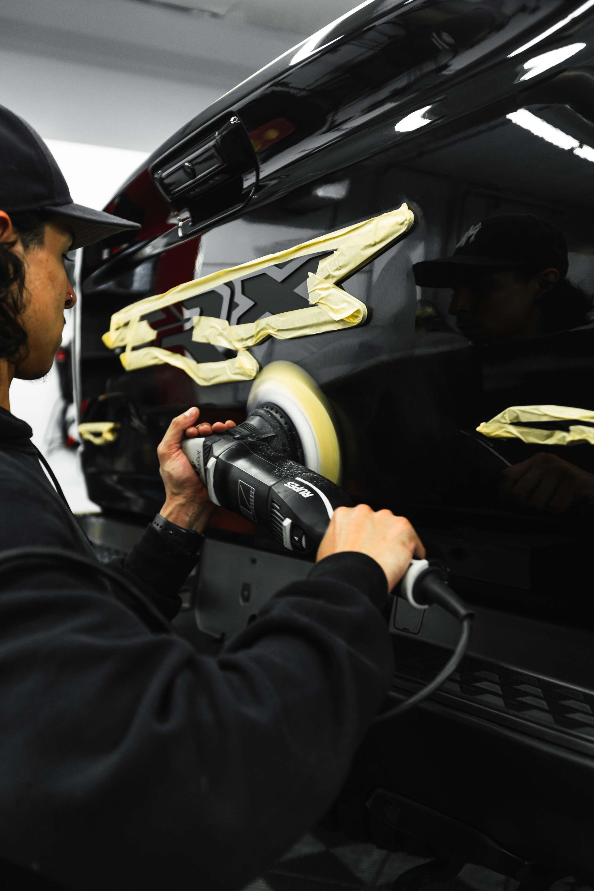 Person polishing a black car door with a buffer, masked areas taped off in a workshop