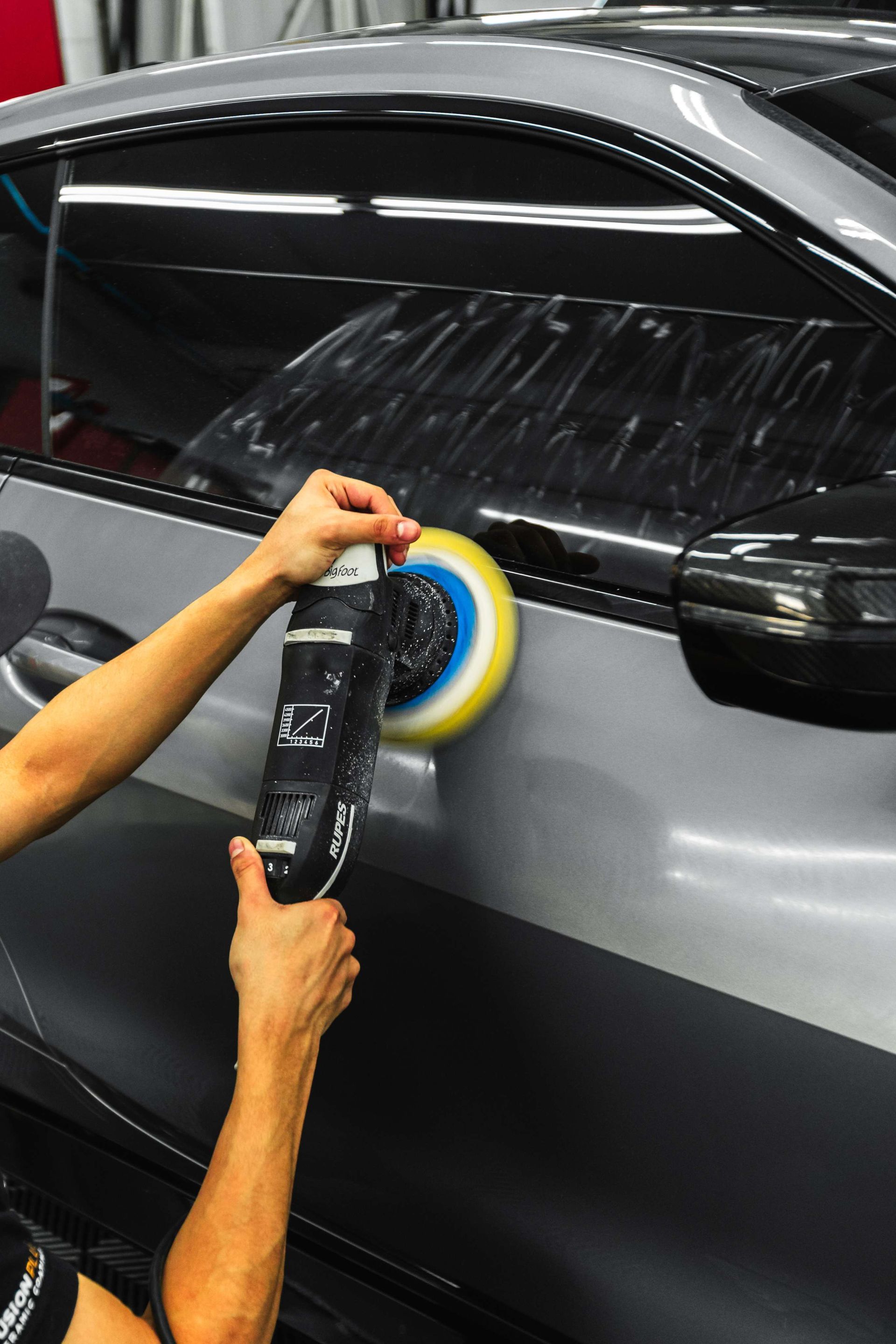 Person buffing a black car door with a polishing machine and yellow-blue pad