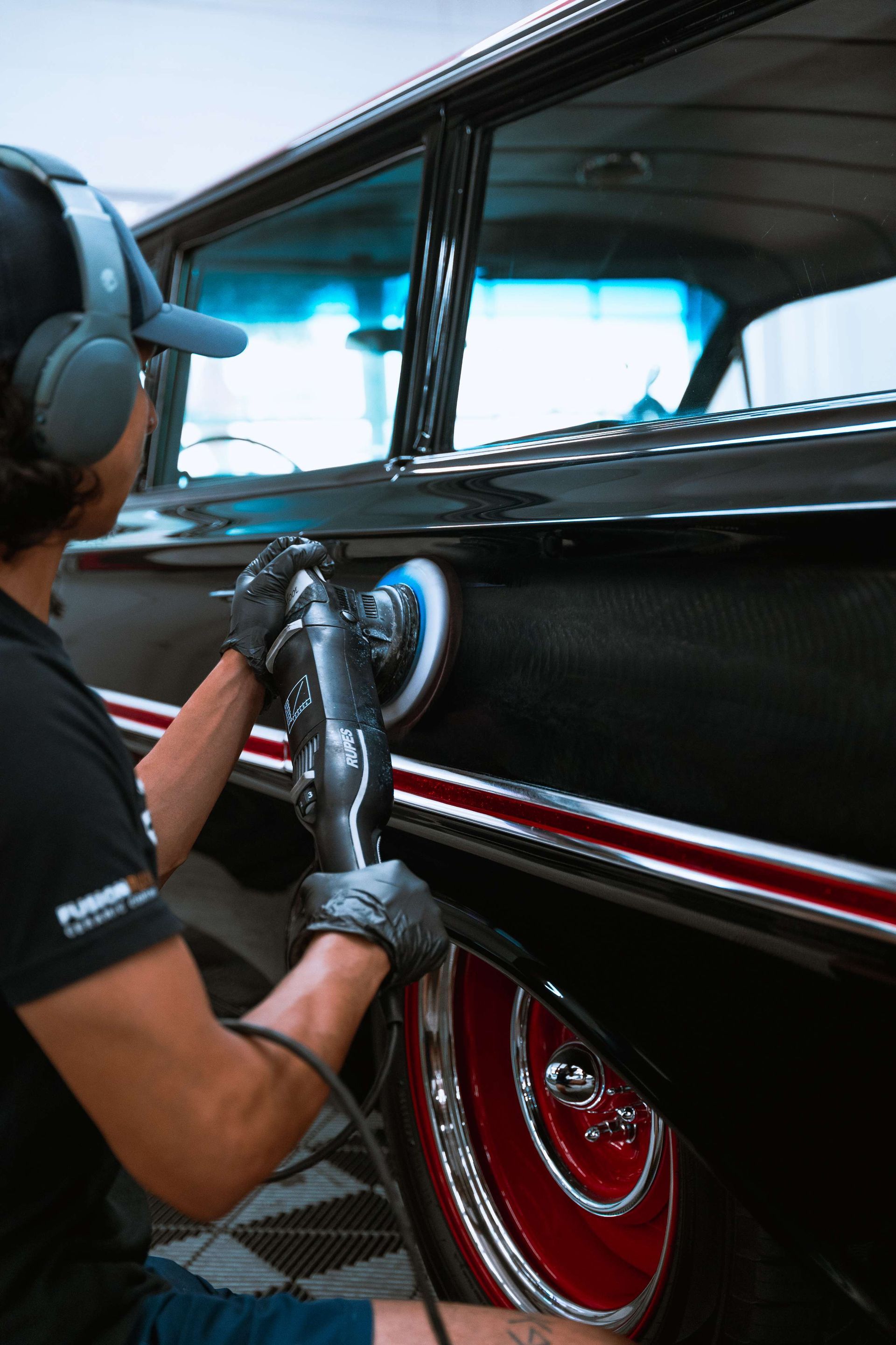 Person polishing a black vintage car with chrome trim and red wheels in a garage