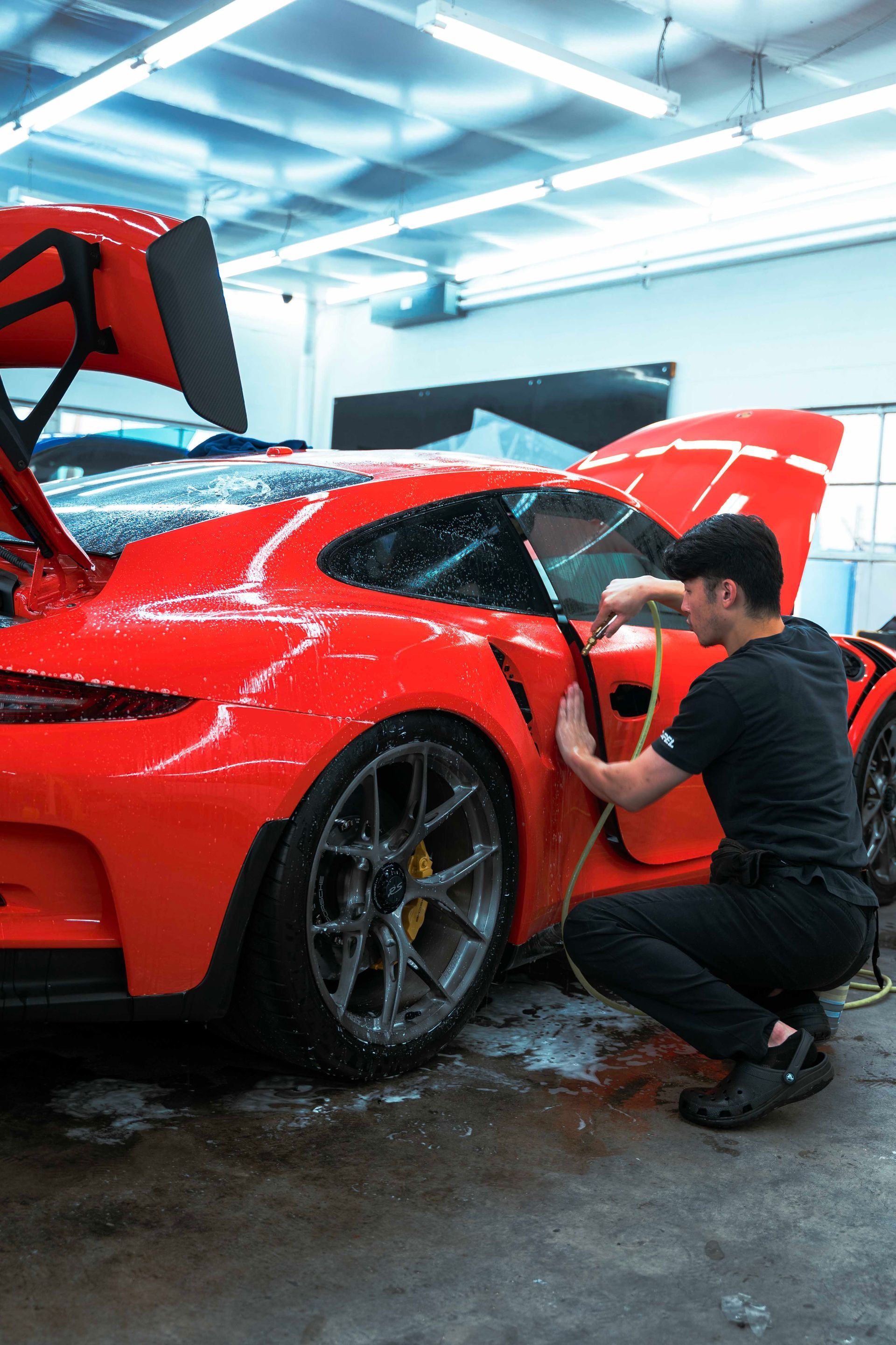 Man washing a red sports car in a garage, with the hood open and water on the floor