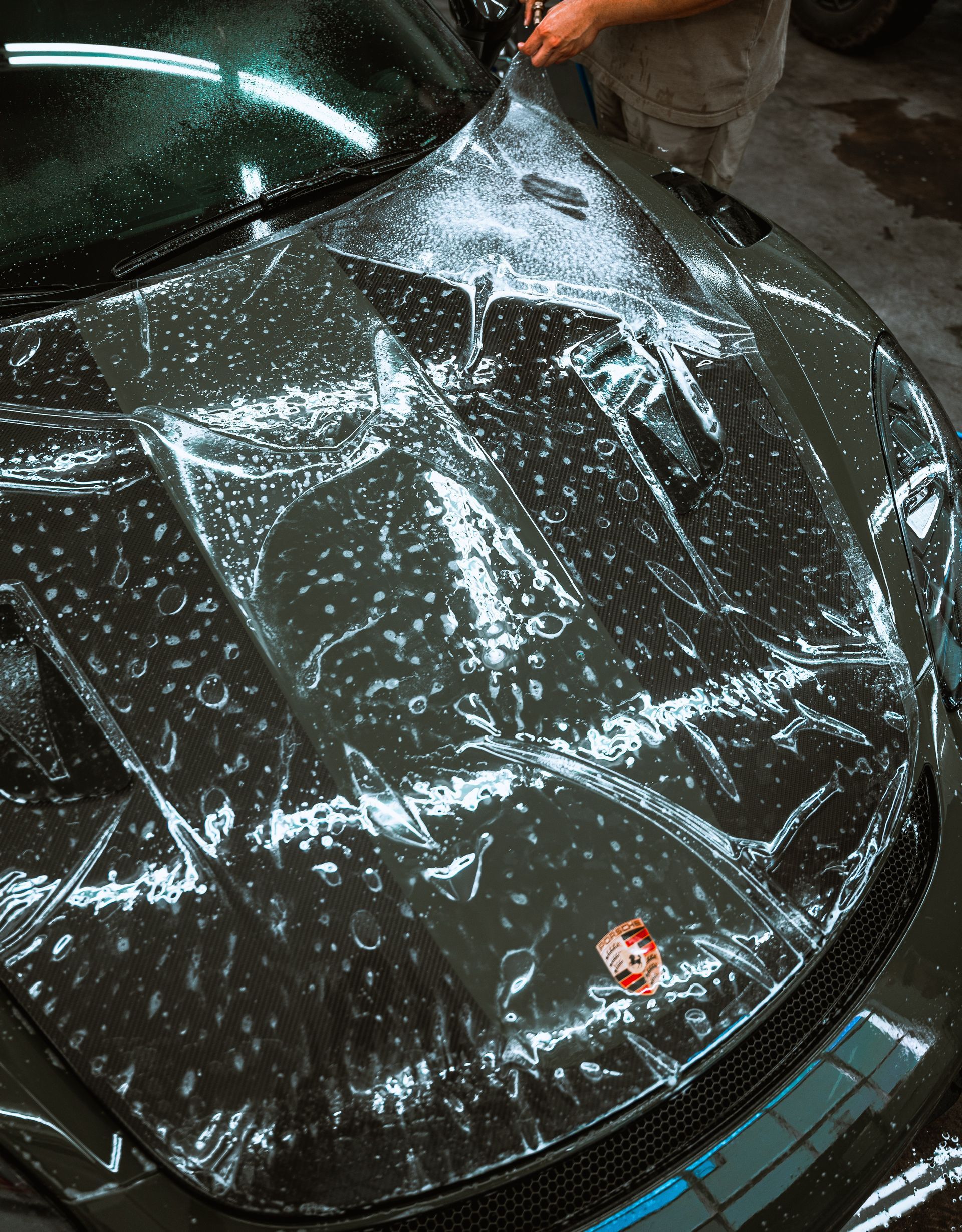 Person applying clear protective film to a blue car bumper in a workshop