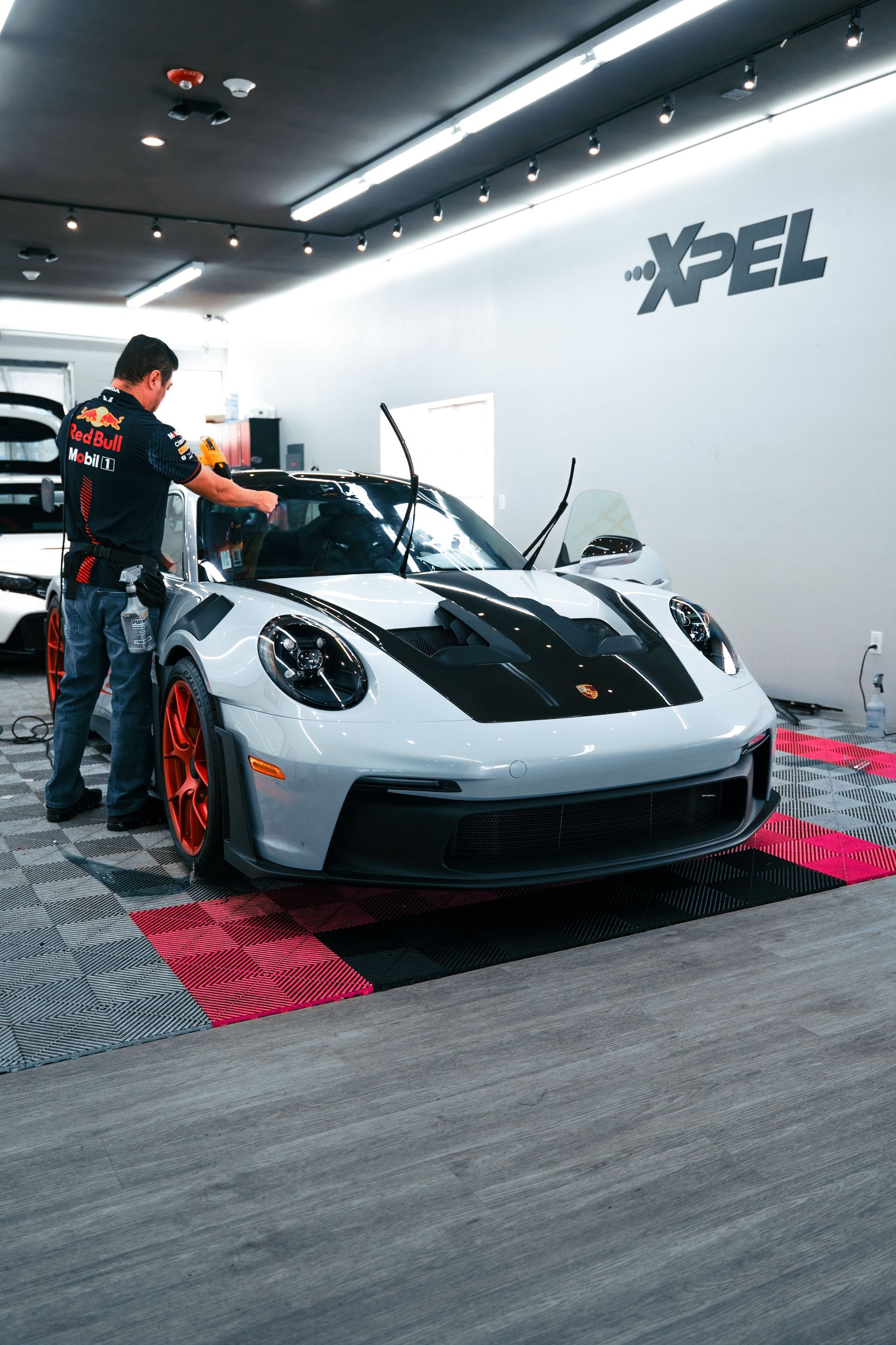 Silver sports car being polished in a bright XPEL garage with black racing stripes.