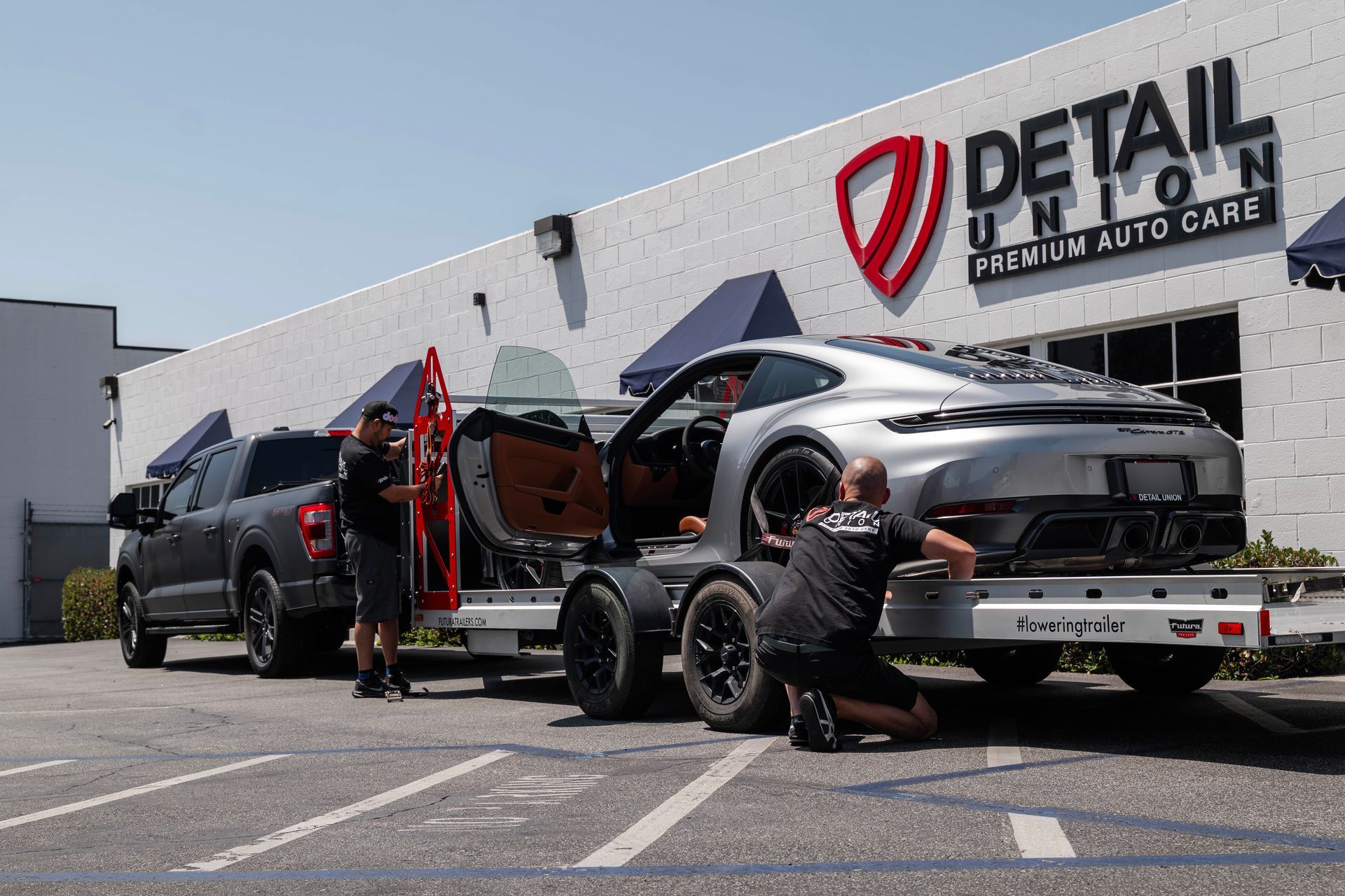 Two people loading a black car onto a trailer outside a Detail automotive care shop