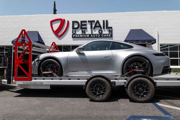Silver sports car on a flatbed trailer outside a detail shop with a white brick wall and sign.