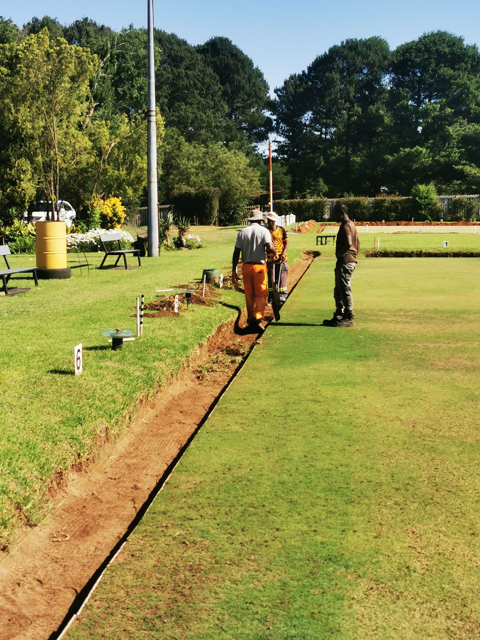 Workers digging a trench beside a grassy field with poles and trees in the background