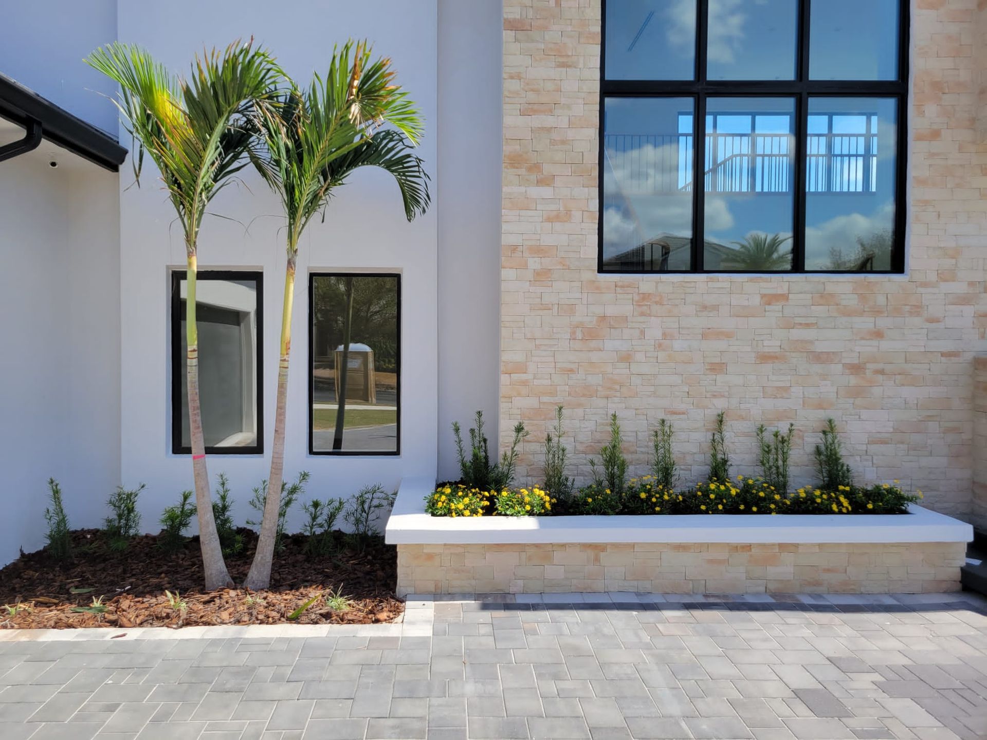 Modern house exterior with palm trees, windows, and brickwork. Landscaping in front of building.