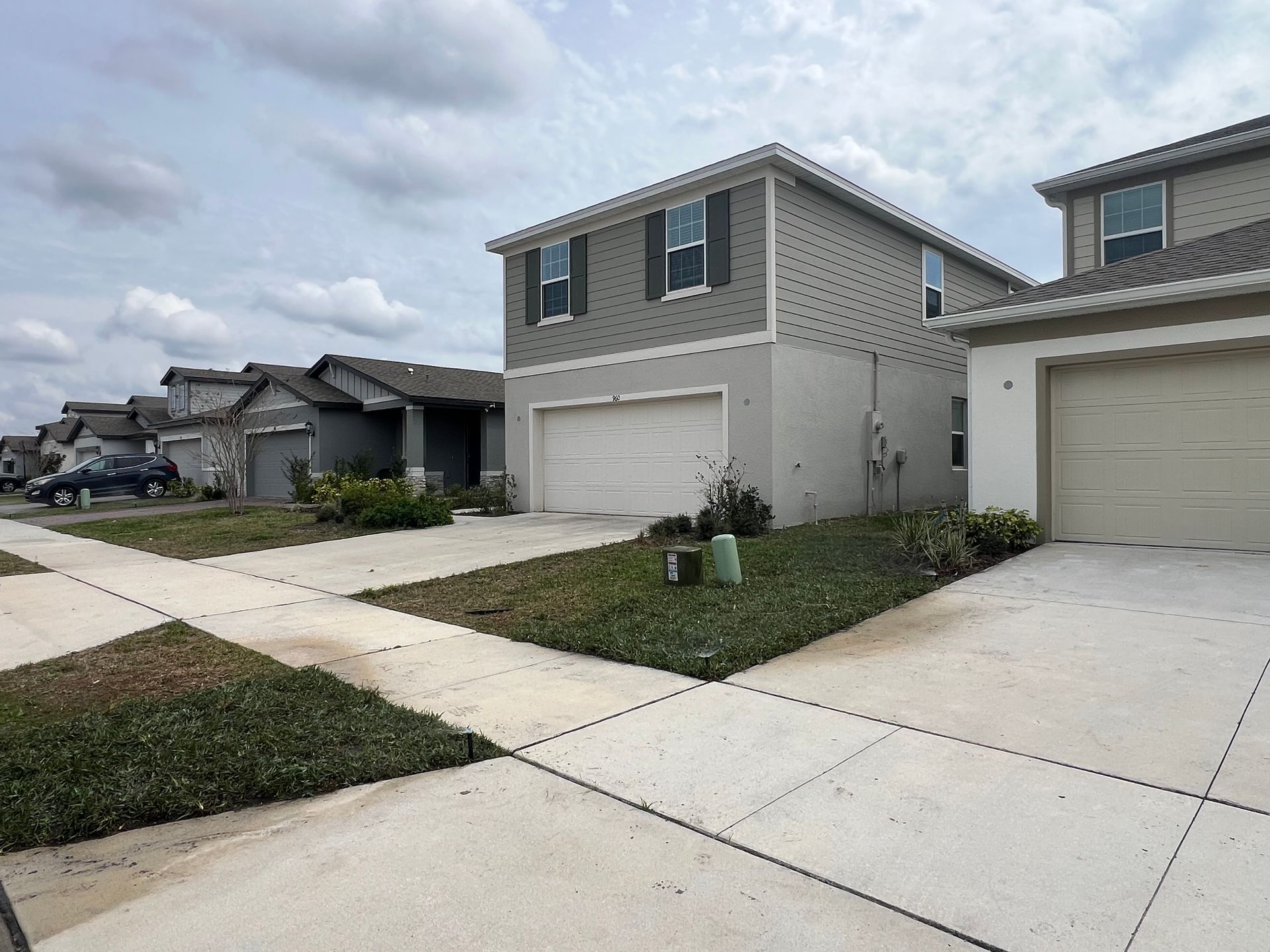 Suburban homes with garages, grey siding, and front lawns along a sidewalk under a cloudy sky.
