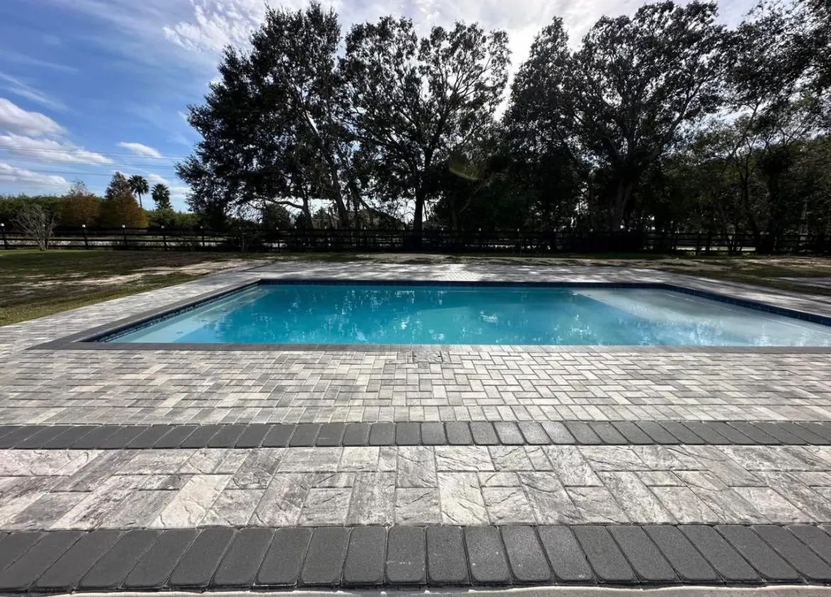 Outdoor rectangular swimming pool with clear blue water, framed by stone paving and trees under a cloudy sky