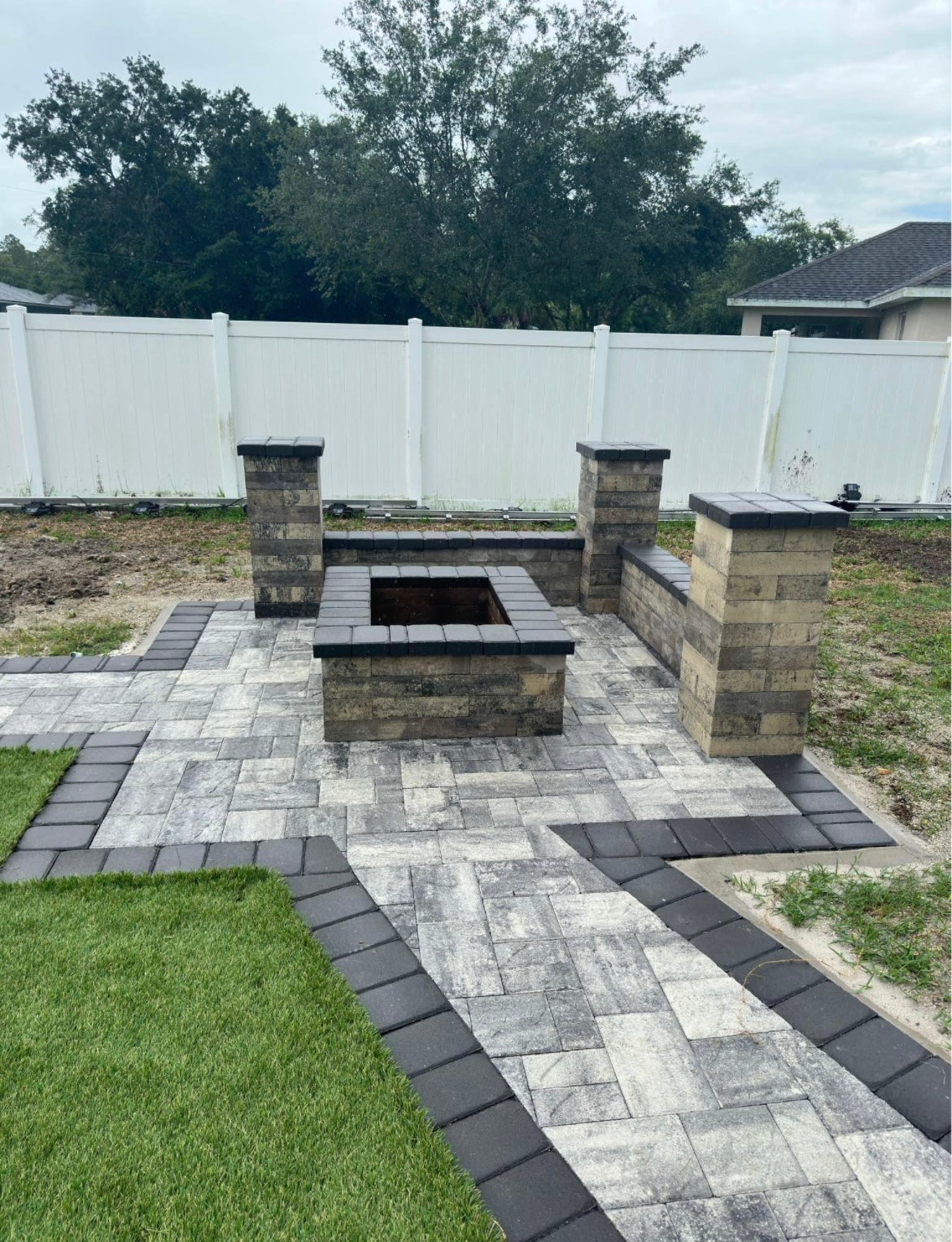 A brick patio with a fire pit, bench, and pillars in a backyard, with a white fence in the background.