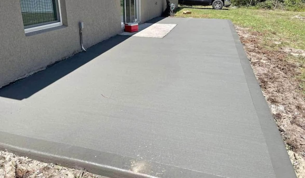 Newly poured concrete patio next to a light gray house; bare dirt surrounds.