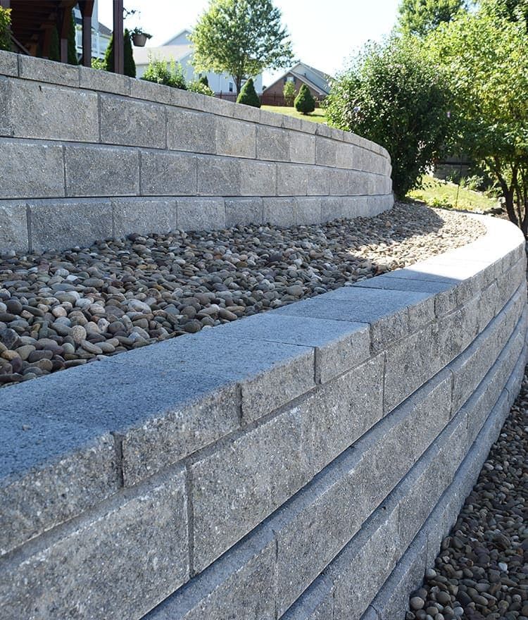 Gray retaining wall curving around a landscaped area with rocks and greenery, sunny outdoor setting.