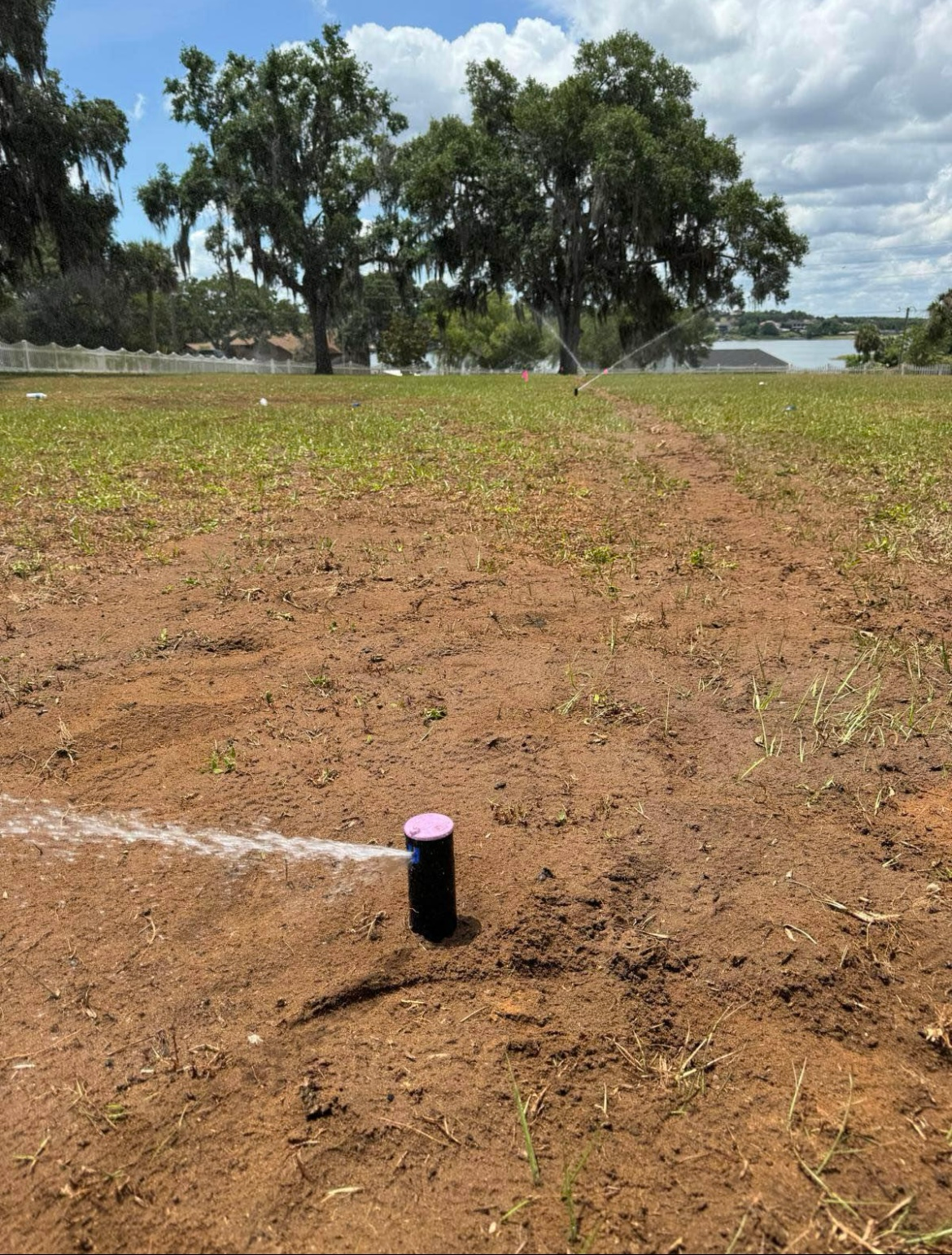 A sprinkler sprays water on dry, brown dirt in a grassy field. Trees and a body of water are in the background.