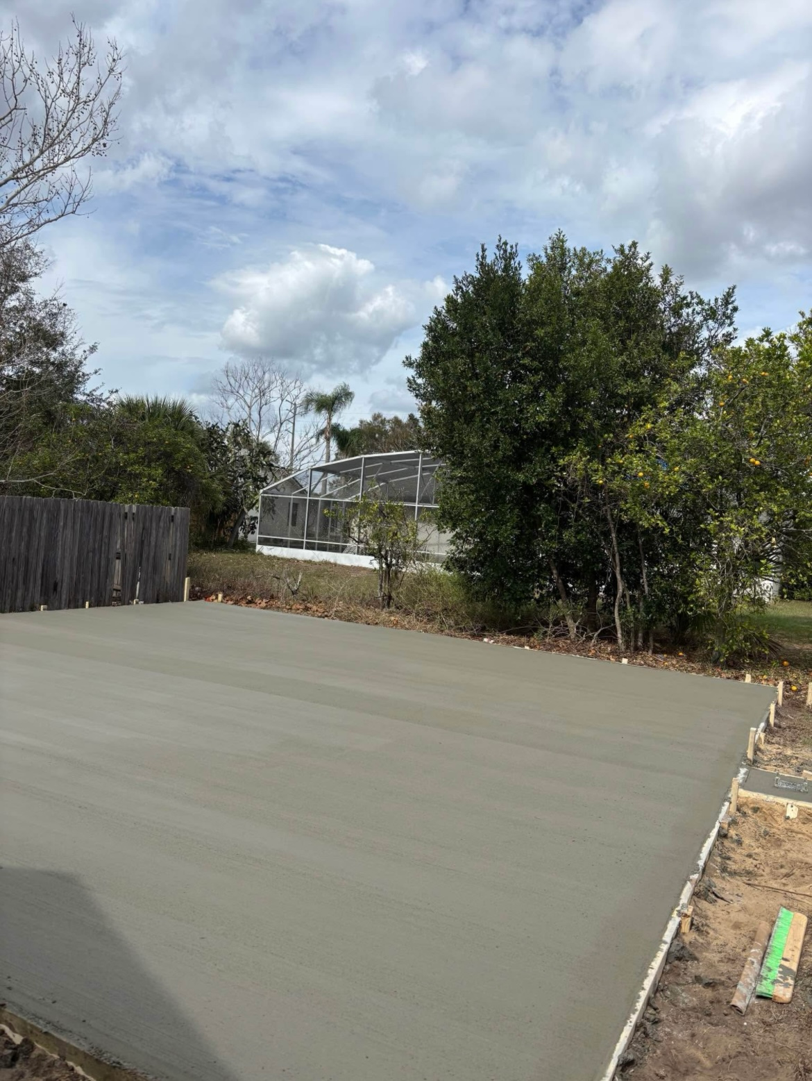 Freshly poured concrete slab outdoors with wooden forms, trees, and a cloudy sky.