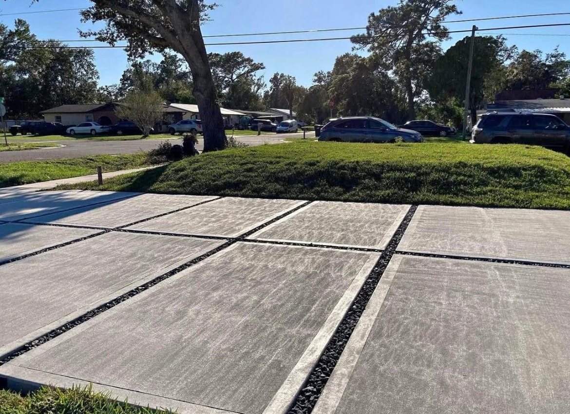 Concrete driveway with decorative gravel-filled lines, green lawn, cars, and houses in the background.