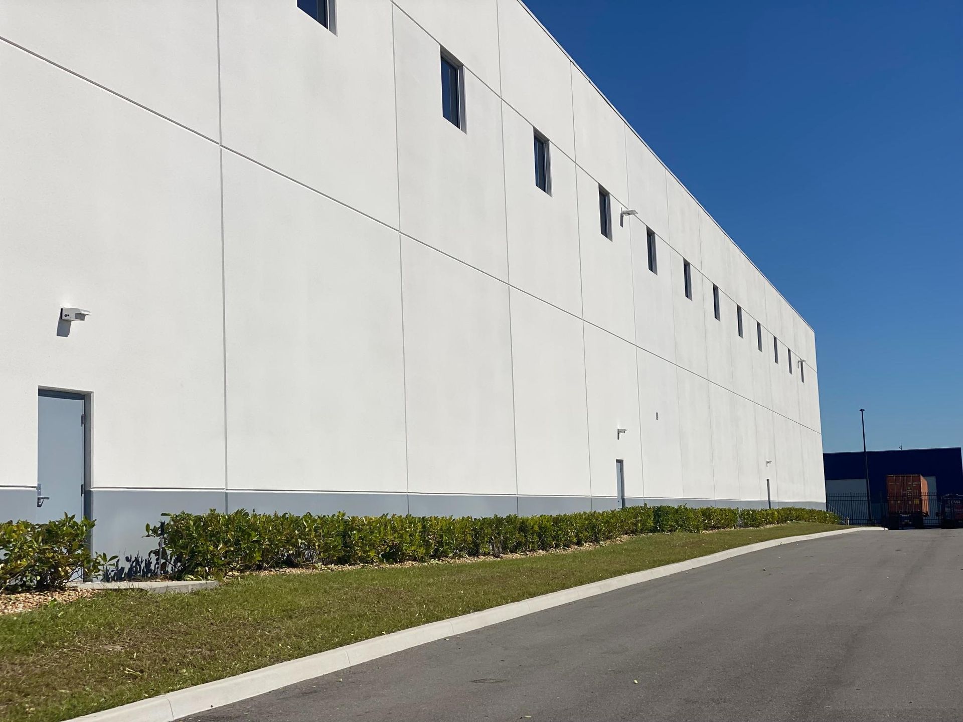 White industrial building exterior with windows, door, and bushes along a dark asphalt road under a blue sky.