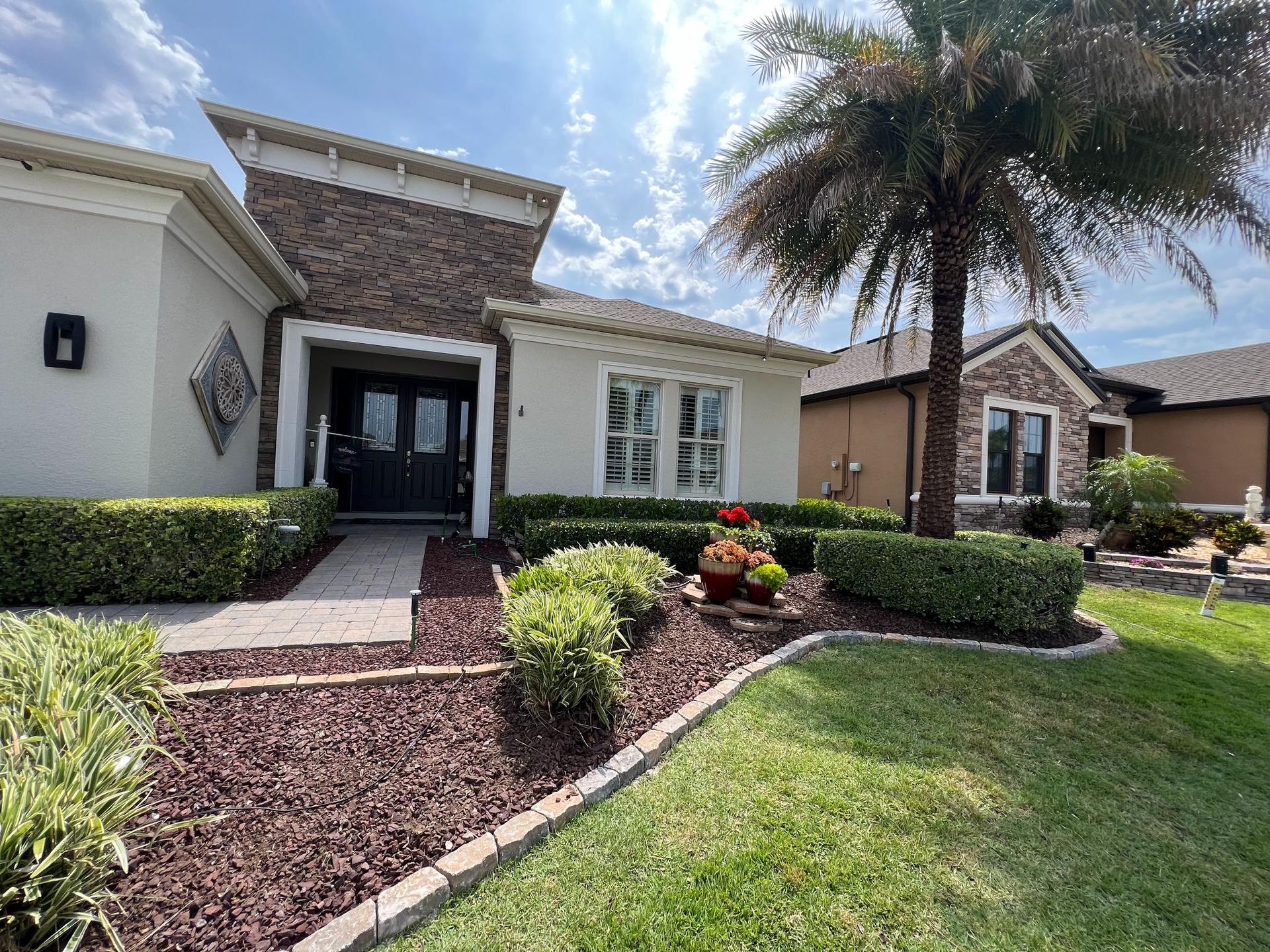 A well-kept house with stone facade above a double front door, surrounded by manicured shrubs and a palm tree.