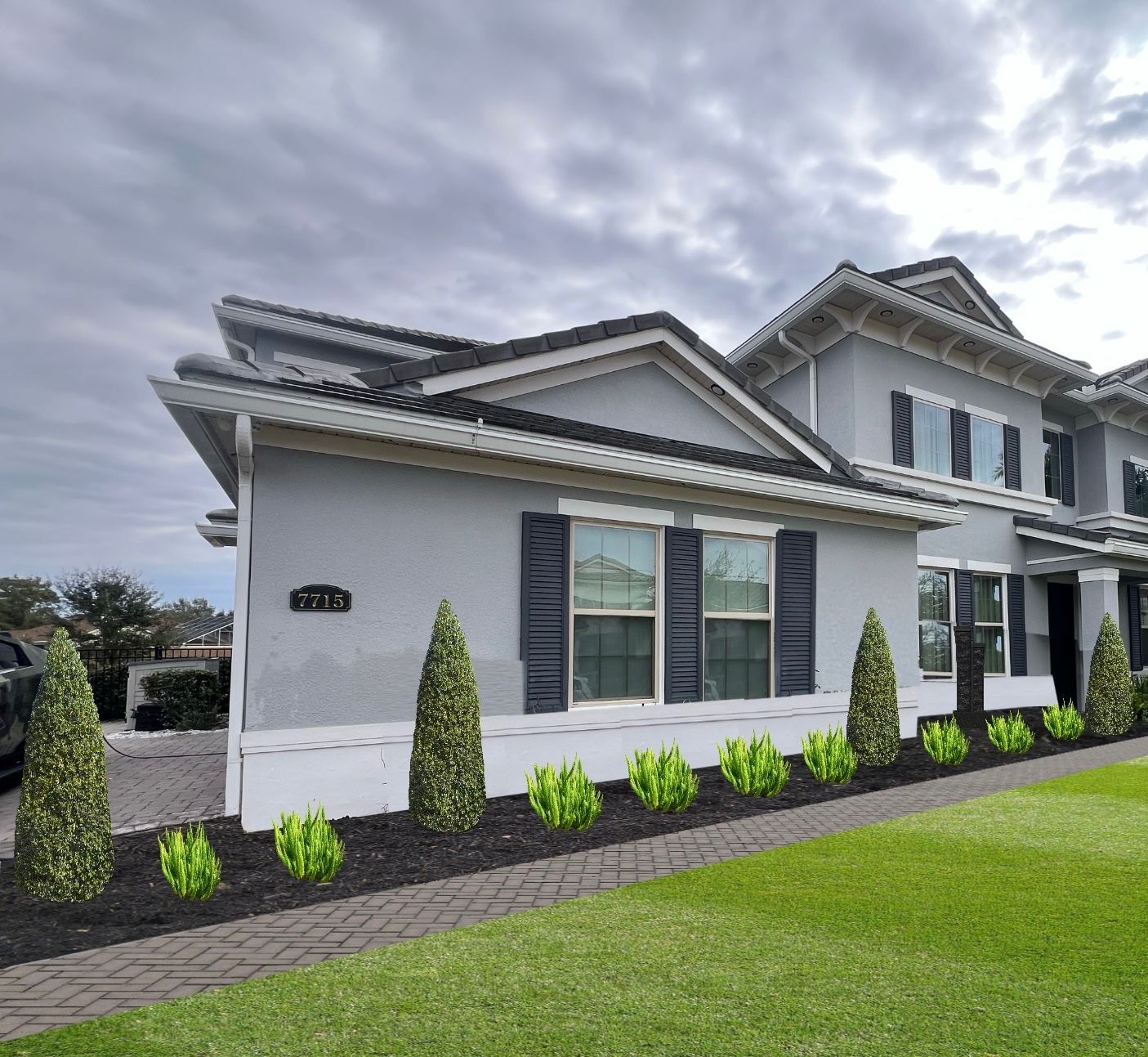Gray stucco house with black shutters and dark roof. Green bushes and grass in front. Overcast sky.