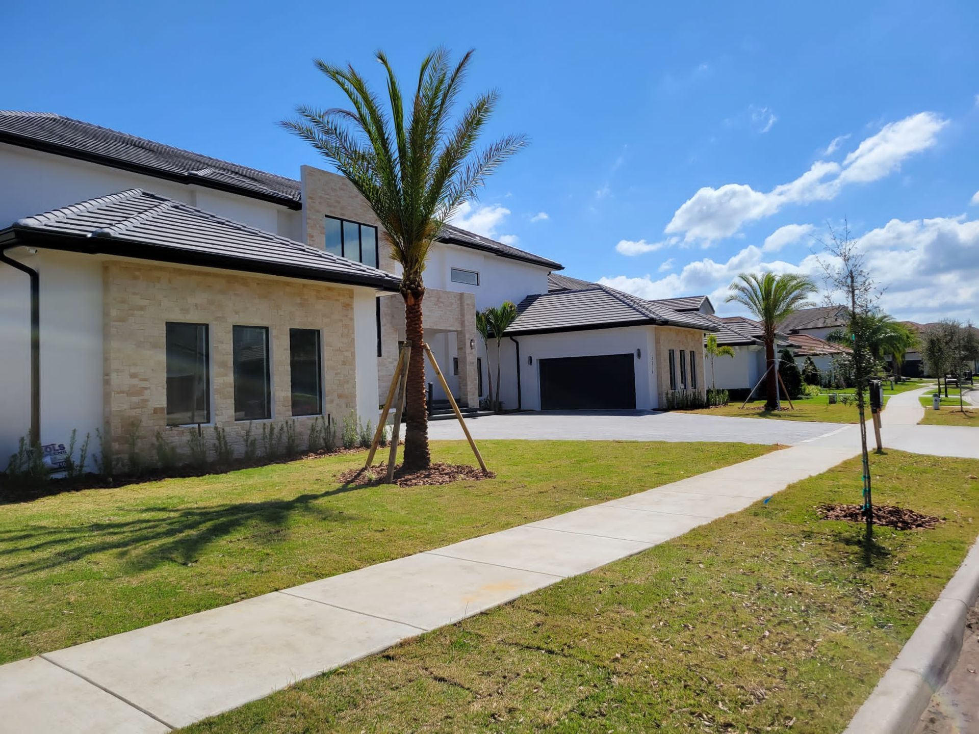 Modern white and stone house with palm trees, driveway, sidewalk, and blue sky.