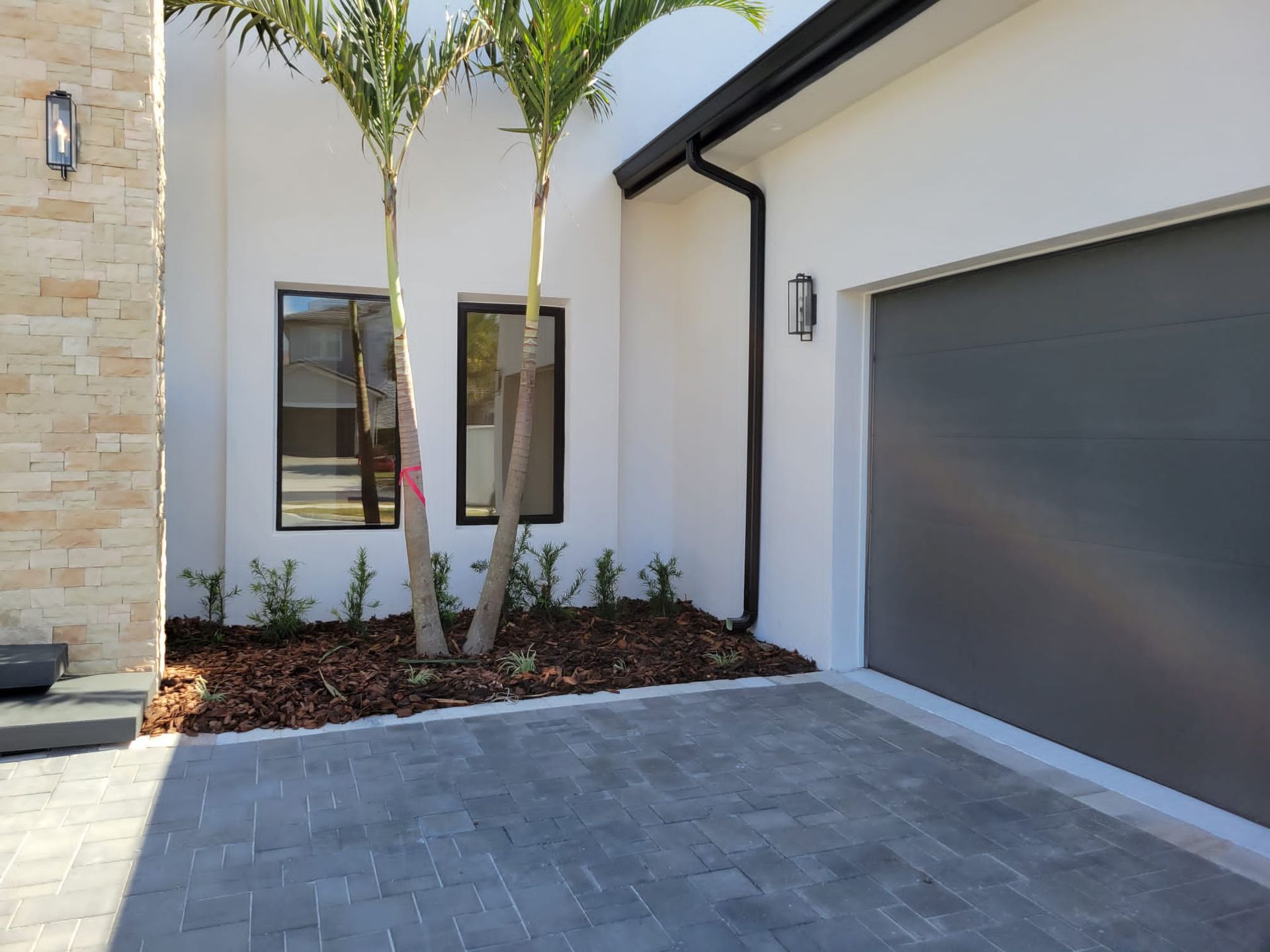 Exterior of a modern home with grey garage door, paved driveway, two palm trees, and white walls.