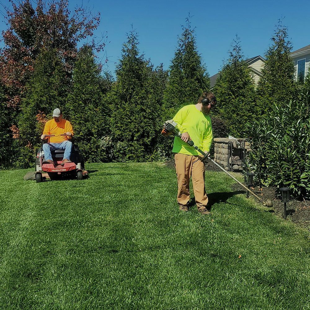 Two people mowing a lush green lawn on a sunny day. One uses a riding mower, the other a string trimmer.