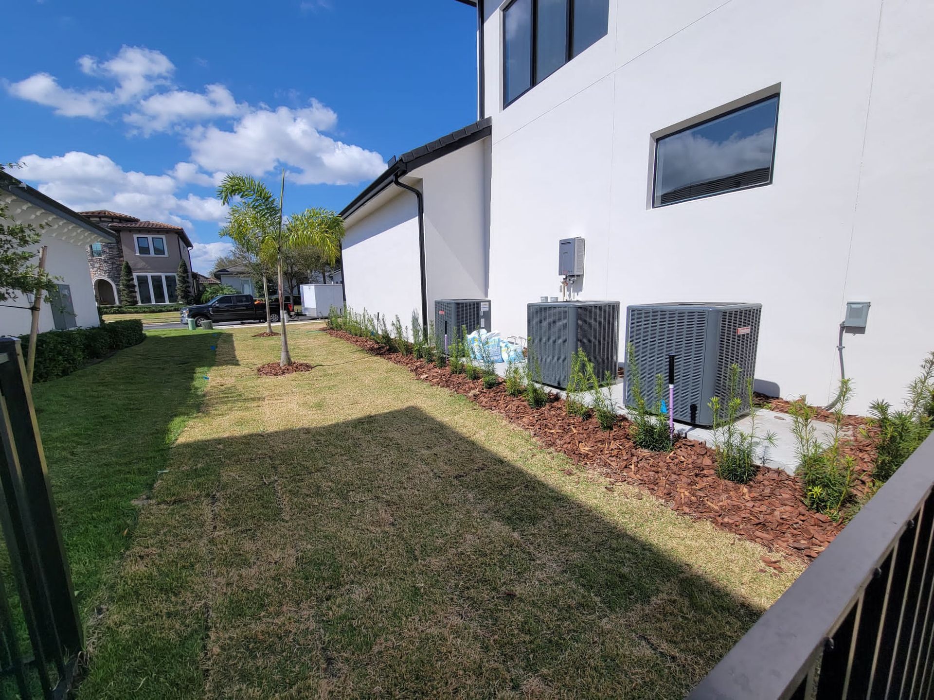 Backyard with lawn, white house, and AC units. Small tree, shrubs, and mulch along the wall. Fence on right, blue sky.