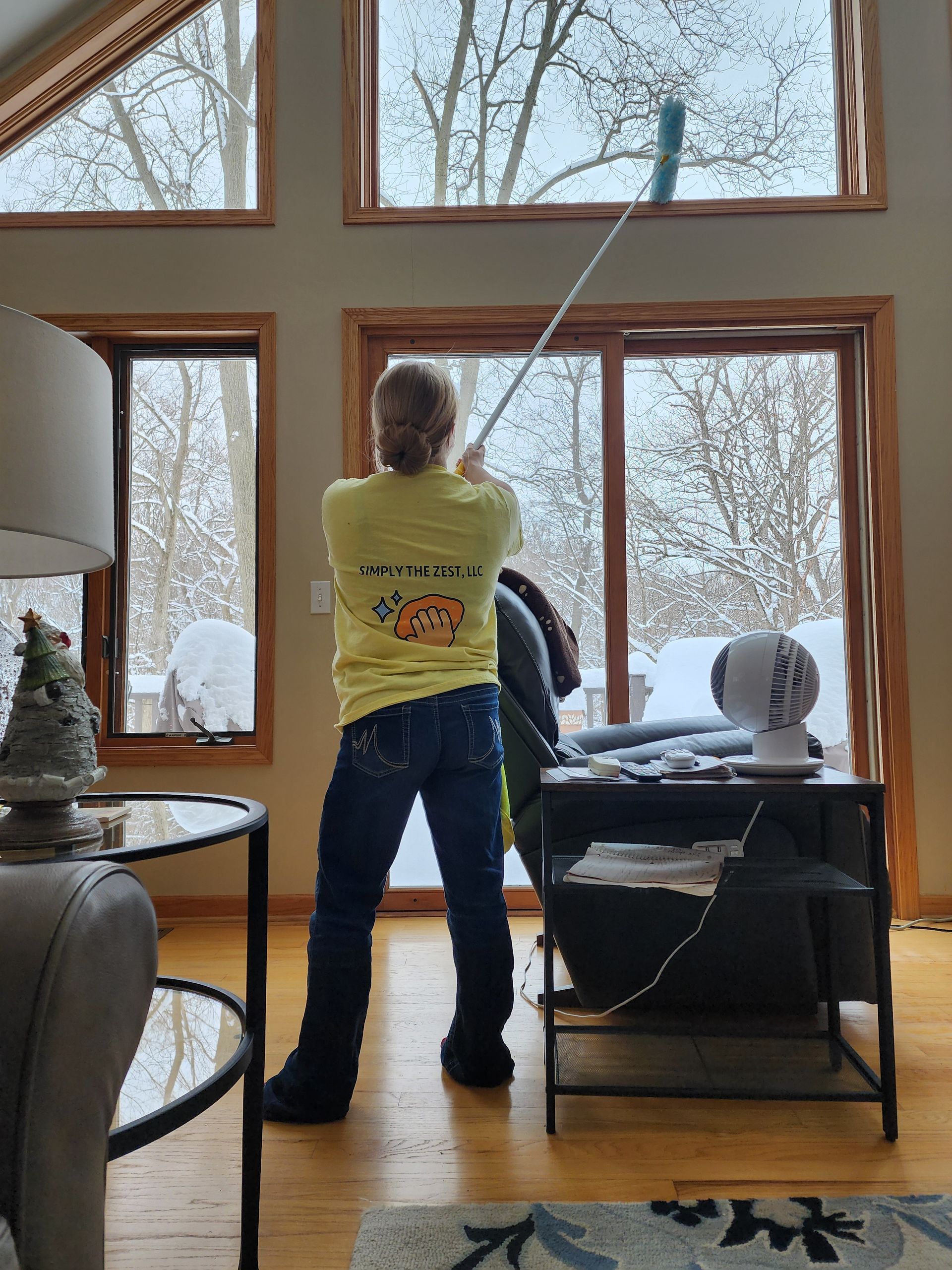 Person cleaning tall window inside, using a long handled squeegee. Snowy outdoor view, light brown trim, wood floors.