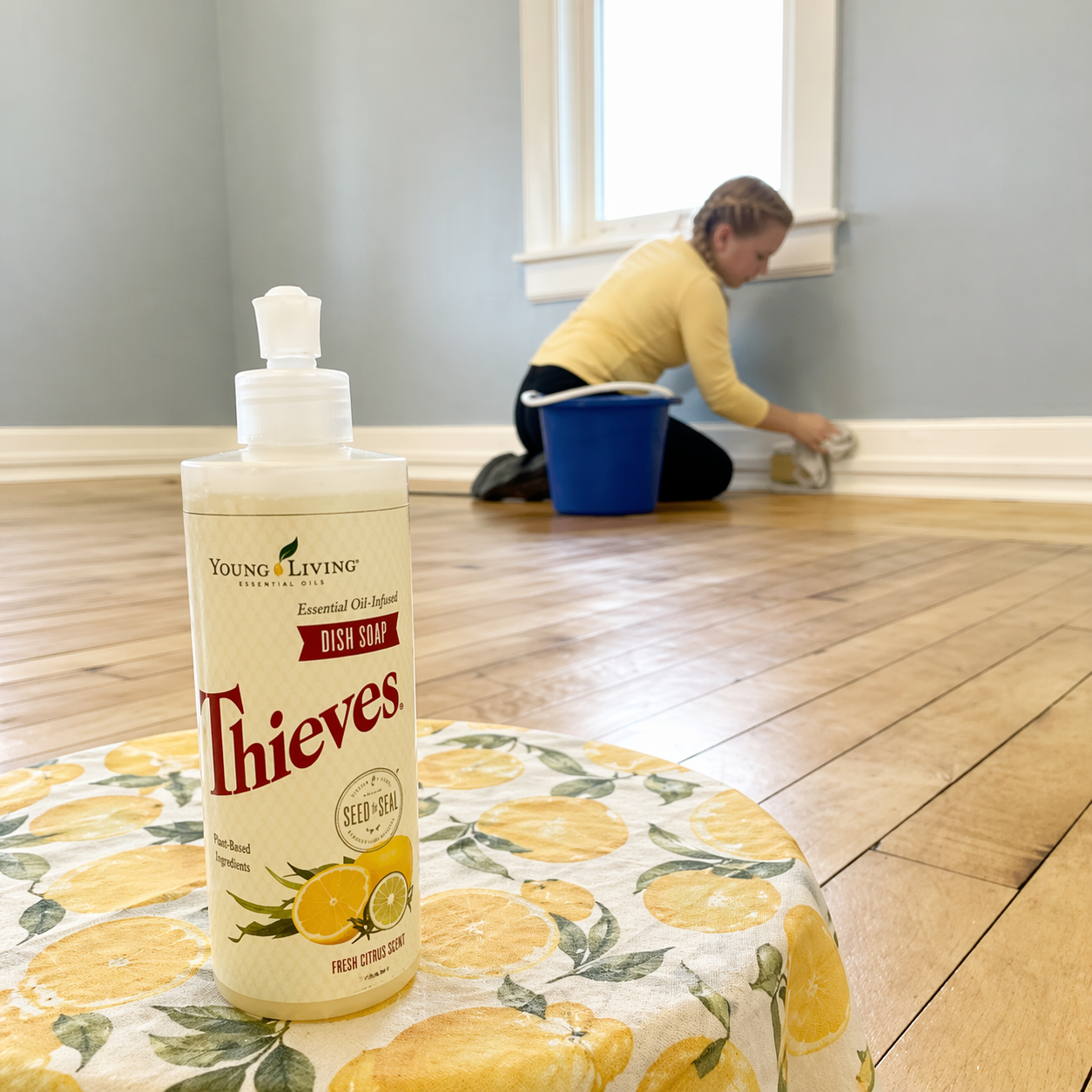 Thieves cleaner bottle on a lemon-patterned cloth in foreground, with a child cleaning a wall in the background.
