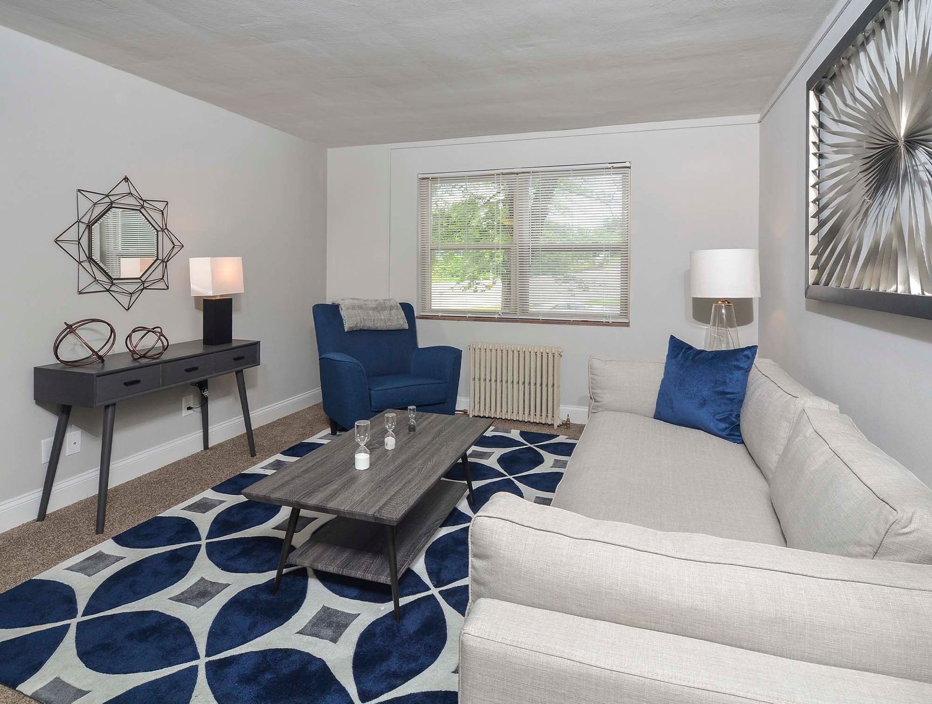 Living room in a modern apartment with a beige sofa, blue armchair, coffee table, and window at The Alden South Hills in Pittsburgh, PA.