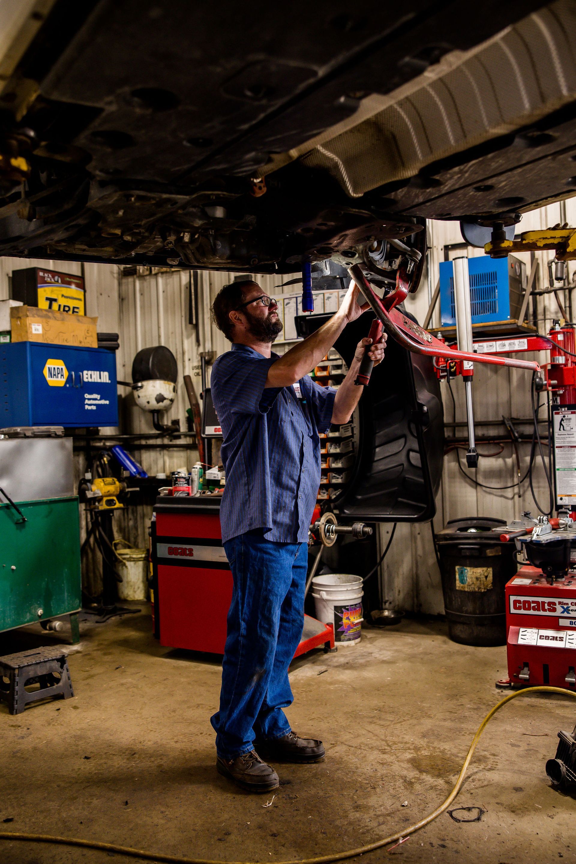 Mechanic working under a car in a garage. Wearing blue work shirt, jeans, and safety glasses.