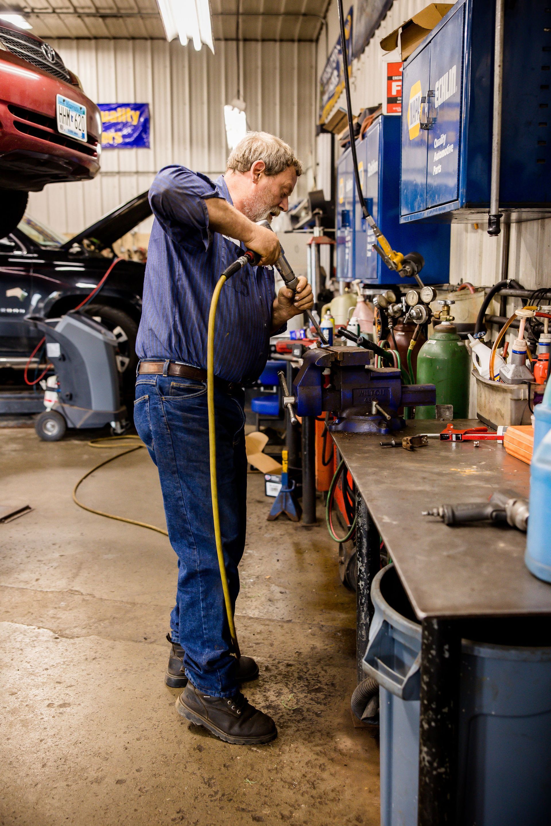 Mechanic uses a tool at a workbench in a cluttered garage, wearing blue shirt and jeans.