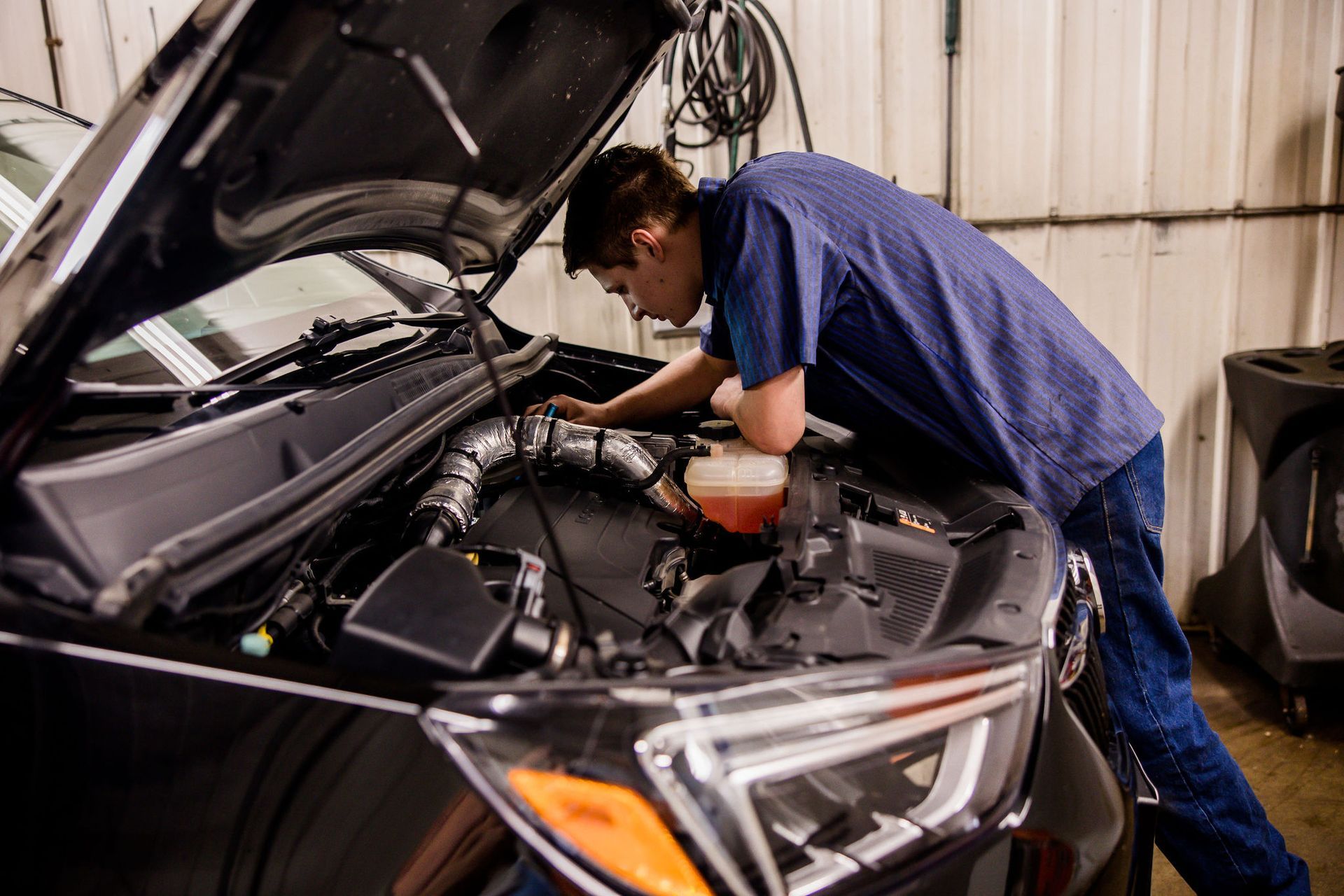 Person working on a car engine in a garage. Hood is open; they are leaning over the engine.