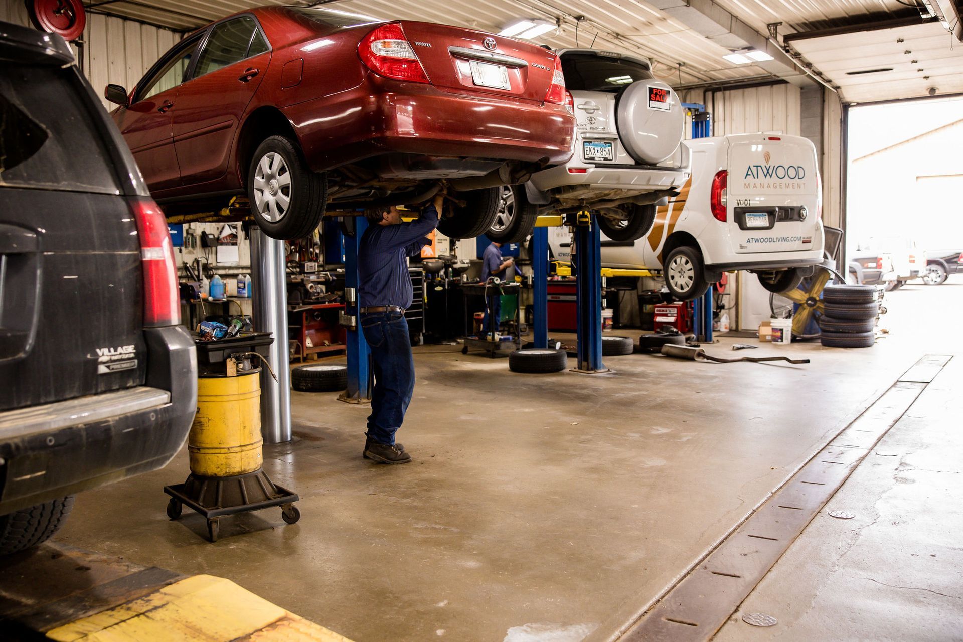 Mechanic working on a red car lifted in an auto repair shop; other vehicles present.