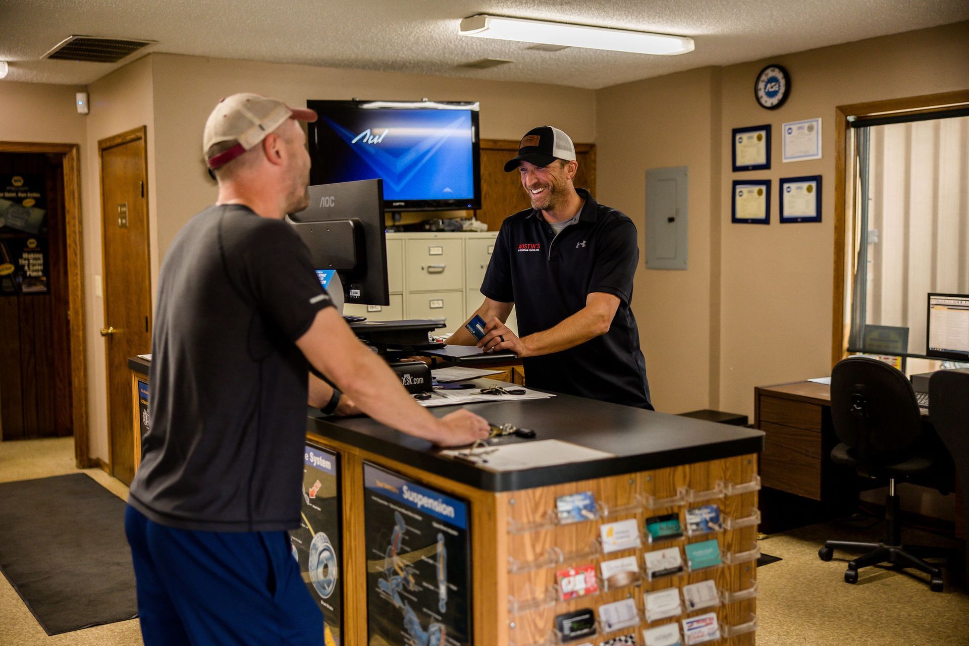 Two men at a reception desk. One is handing the other a card. Office setting with computer and window.