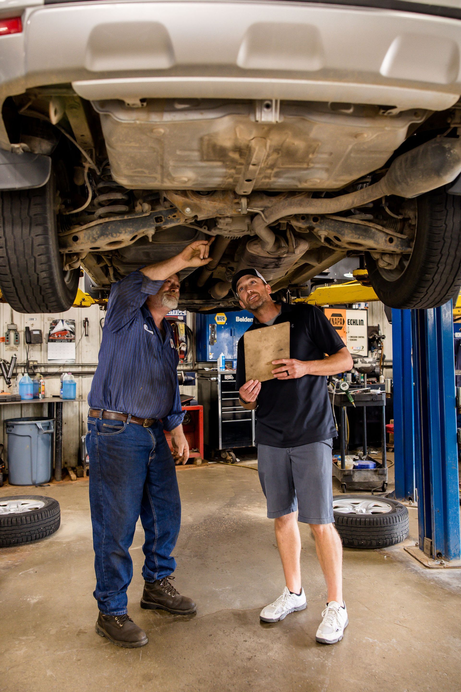 Two men inspecting the underside of a car in a repair shop. One points, the other holds a clipboard.