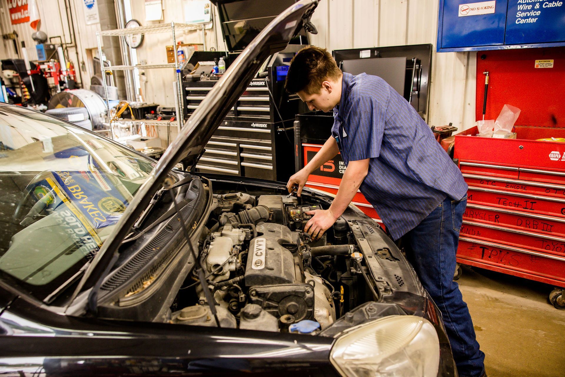 Mechanic working on car engine in a shop, wearing blue shirt and jeans. Hood open.