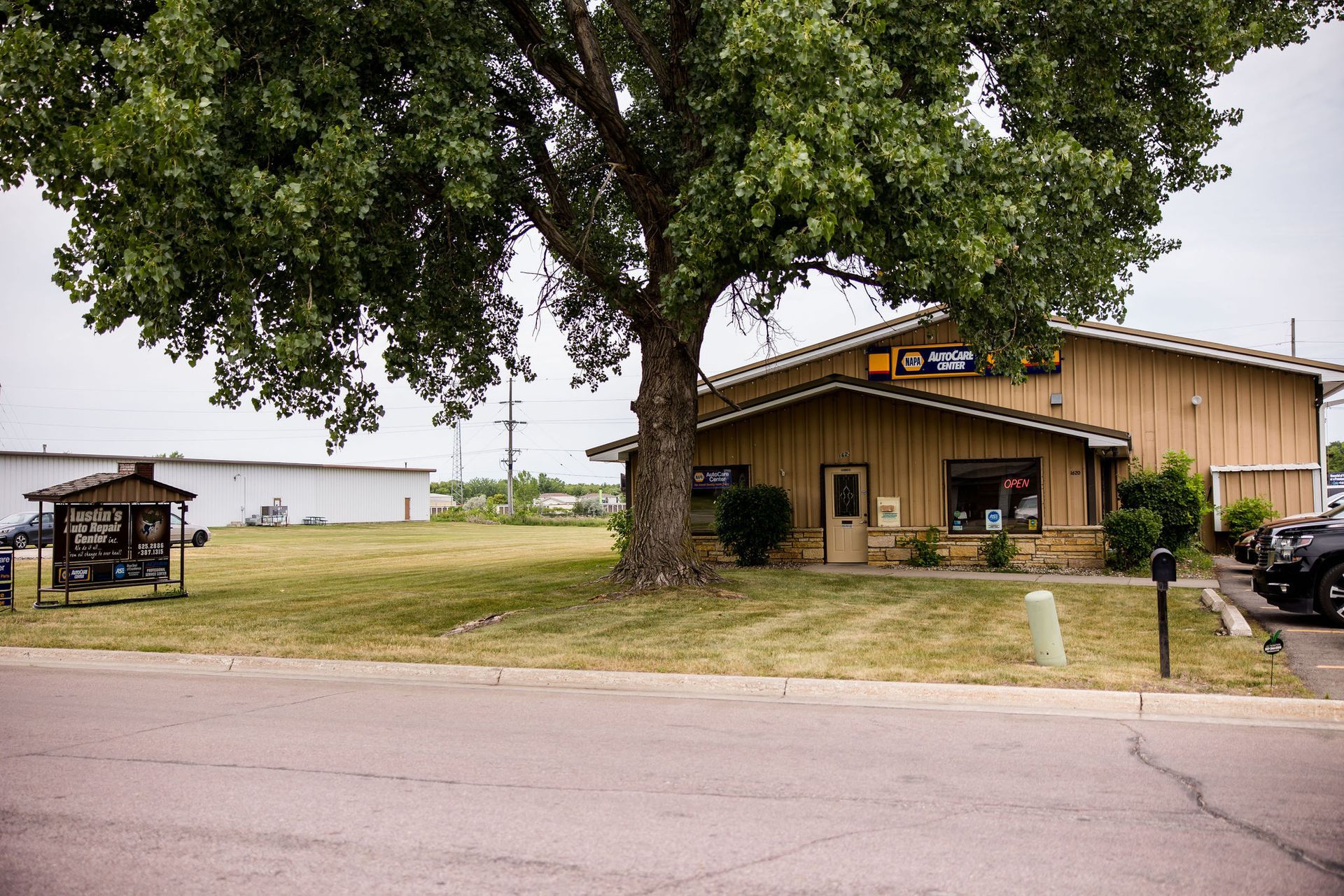 A tan-colored building with a large tree in front; a grassy lawn and a paved street.