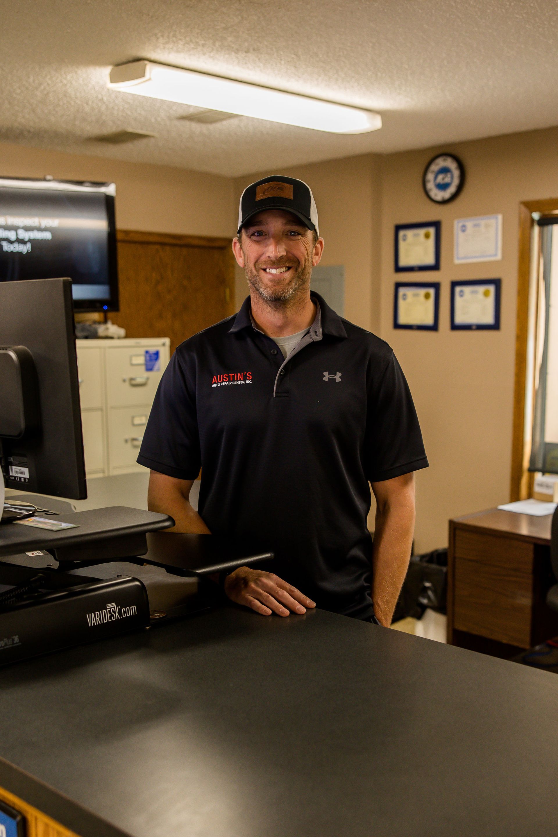 Man in black shirt and hat, smiling, behind a counter in an office setting.