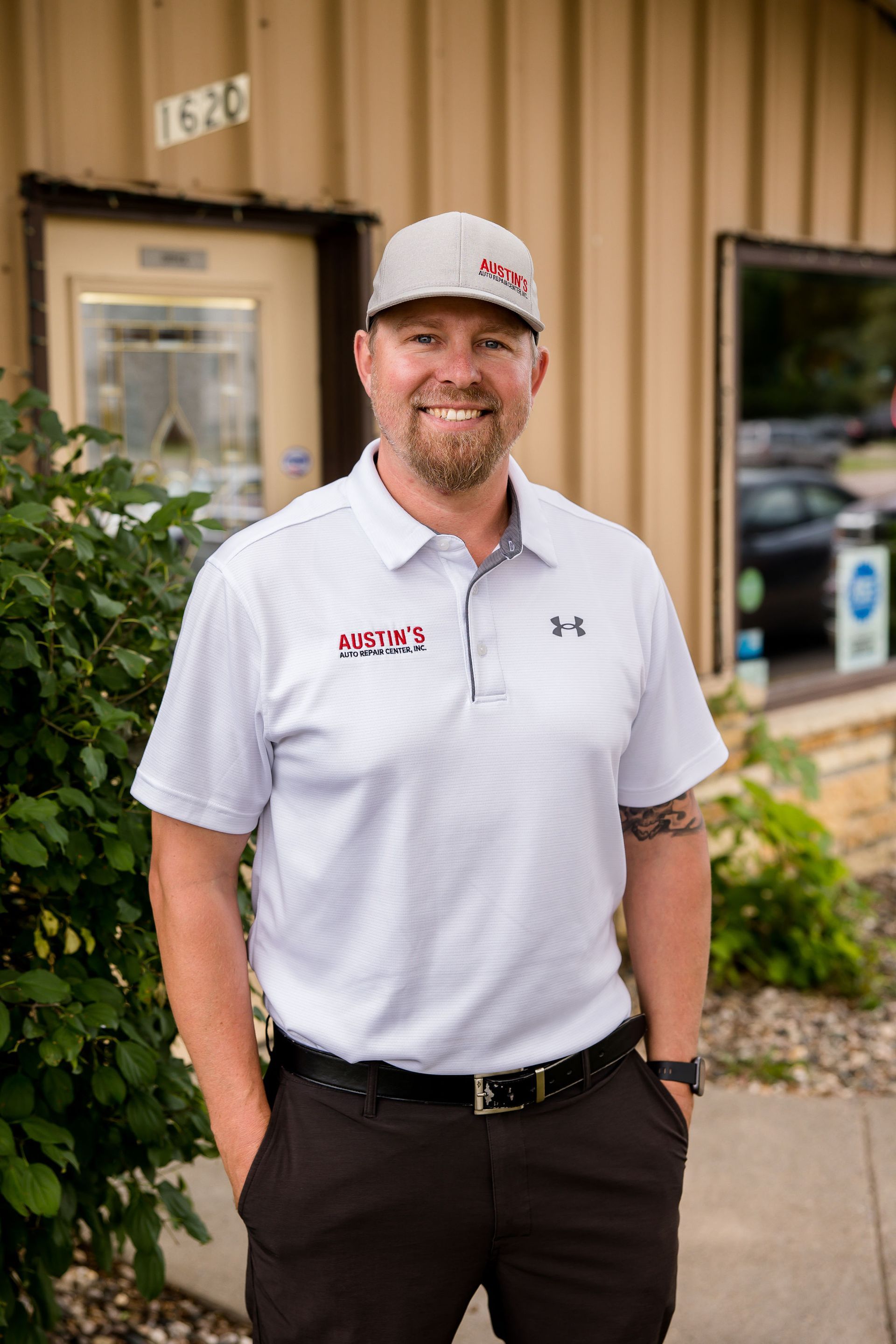 Man wearing a hat and polo shirt stands in front of a building with his hands in pockets.