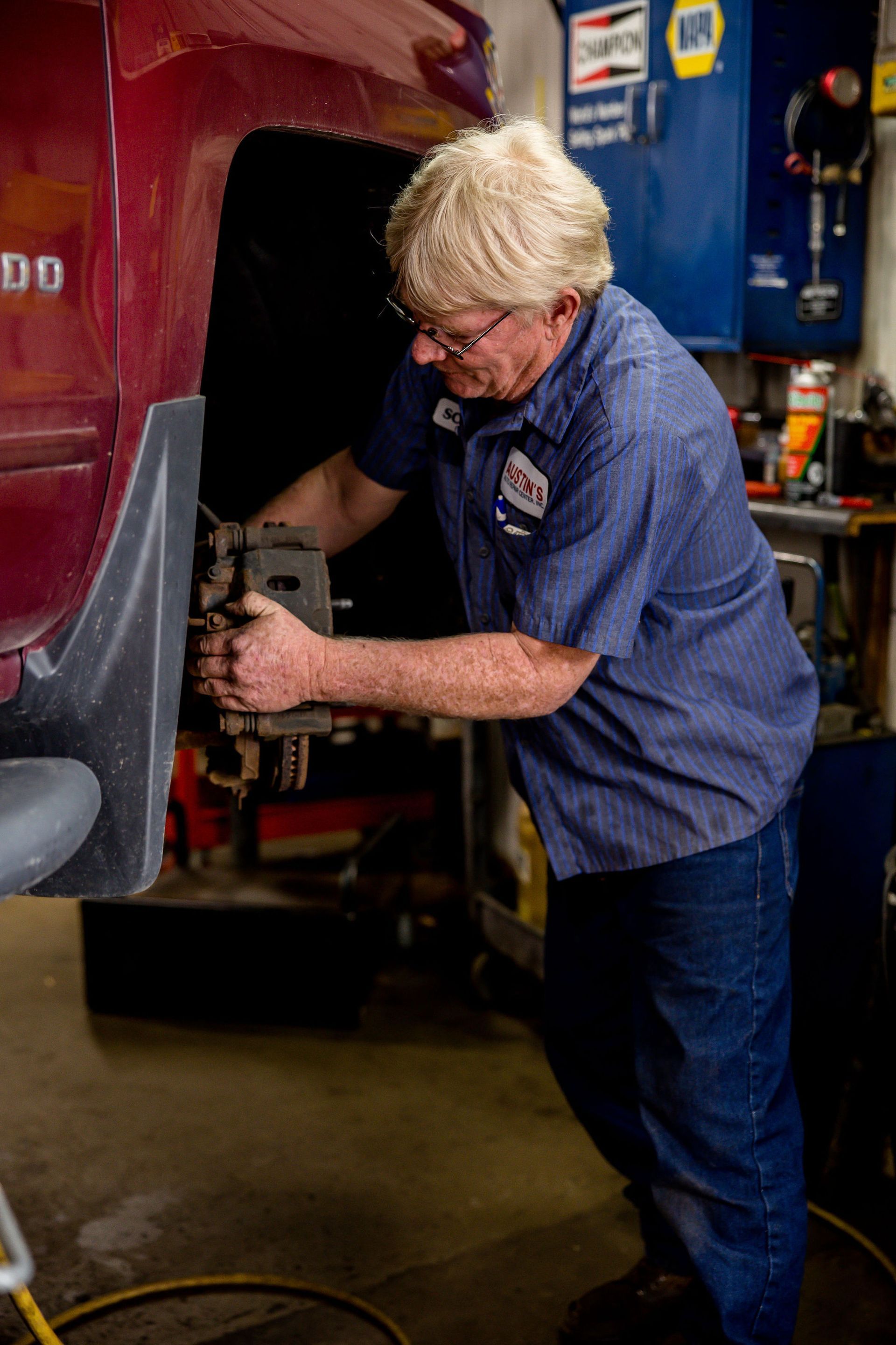 Mechanic working on the brakes of a red vehicle in a garage. Wearing blue work shirt, jeans, and glasses.