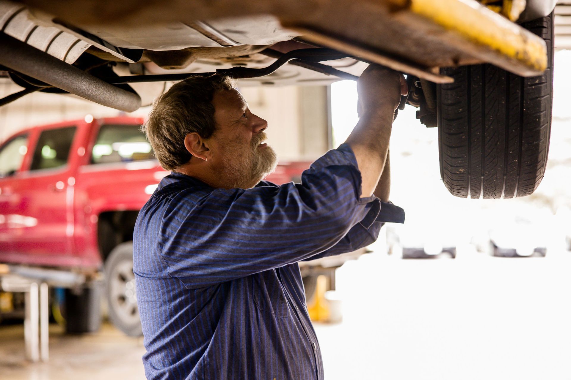 Mechanic working on the underside of a car in a garage, a red truck in the background.