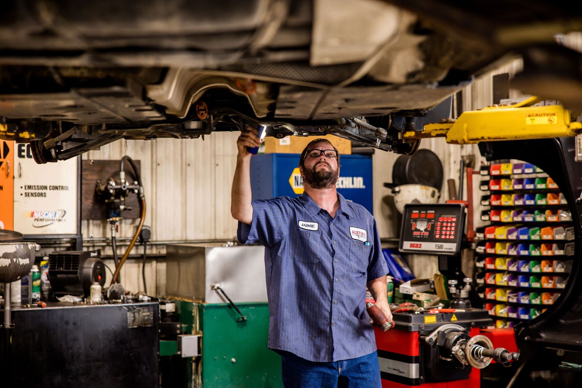 Mechanic examining undercarriage of a car with a flashlight in a garage.