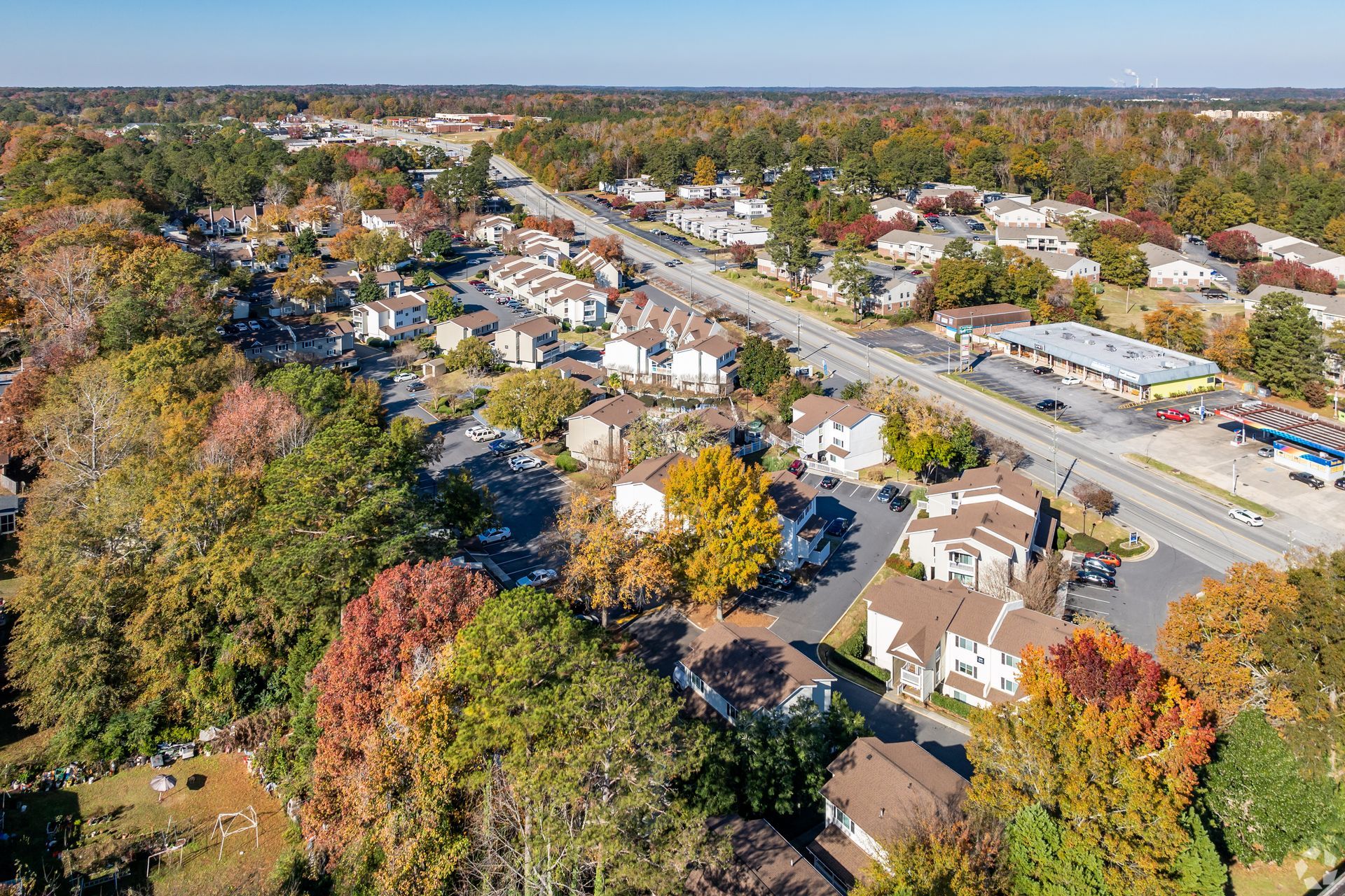 Aerial view of suburban homes and a shopping center surrounded by autumn trees and a road under a clear blue sky.
