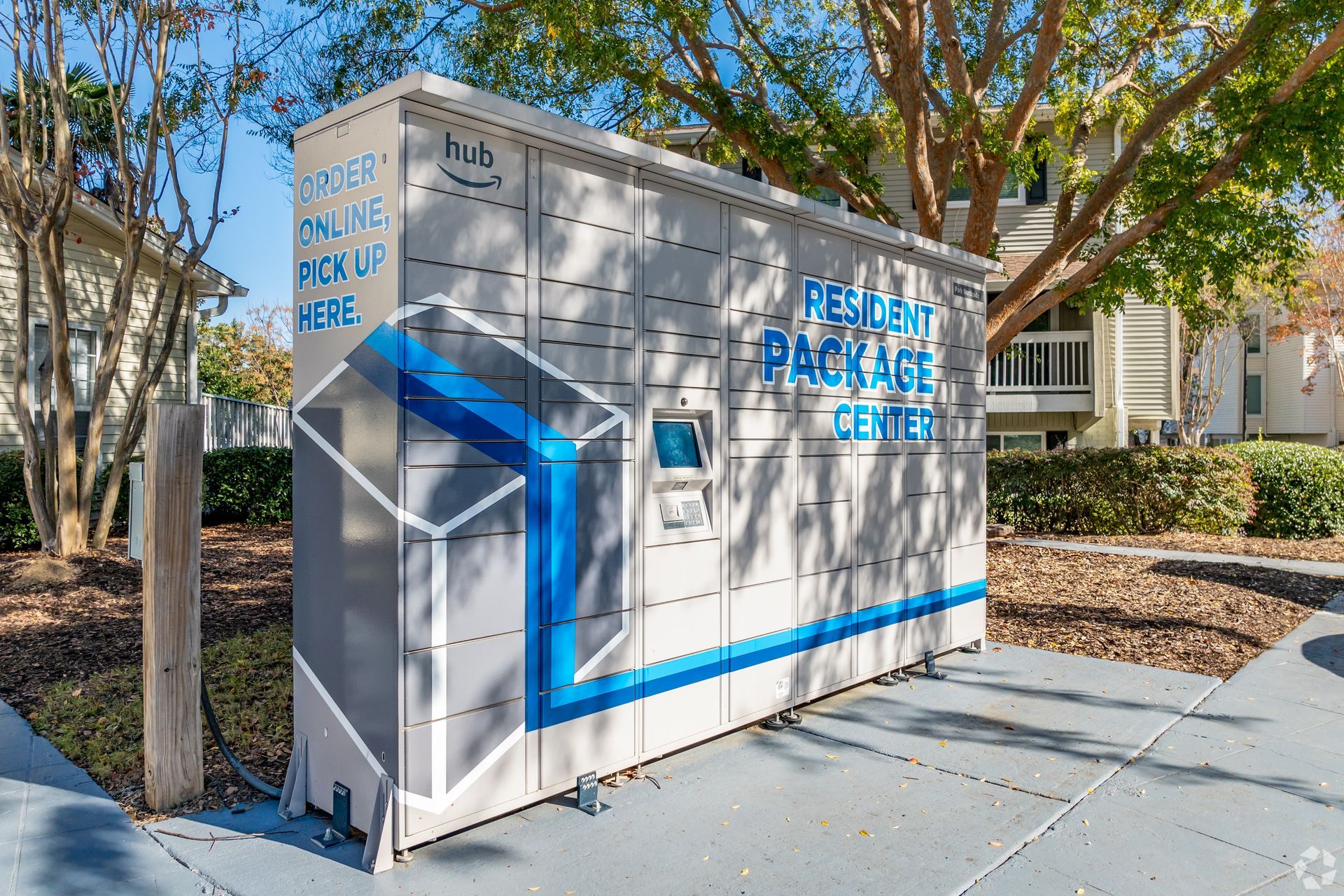 An Amazon Hub package locker in a sunny outdoor setting with trees and apartment buildings in the background.
