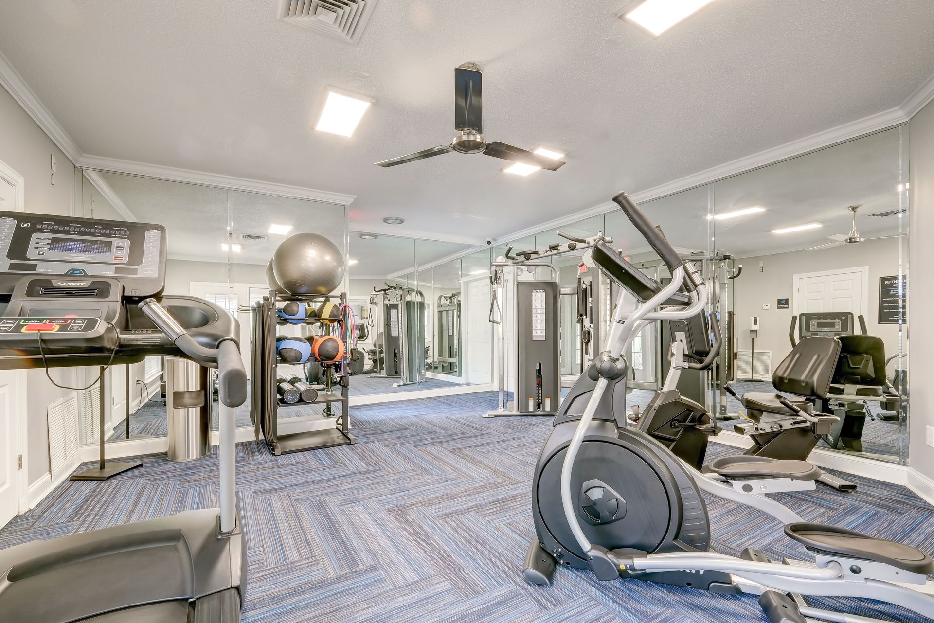 A gym with a treadmill, elliptical machine, and rack of exercise balls on a blue patterned carpet in a mirrored room.