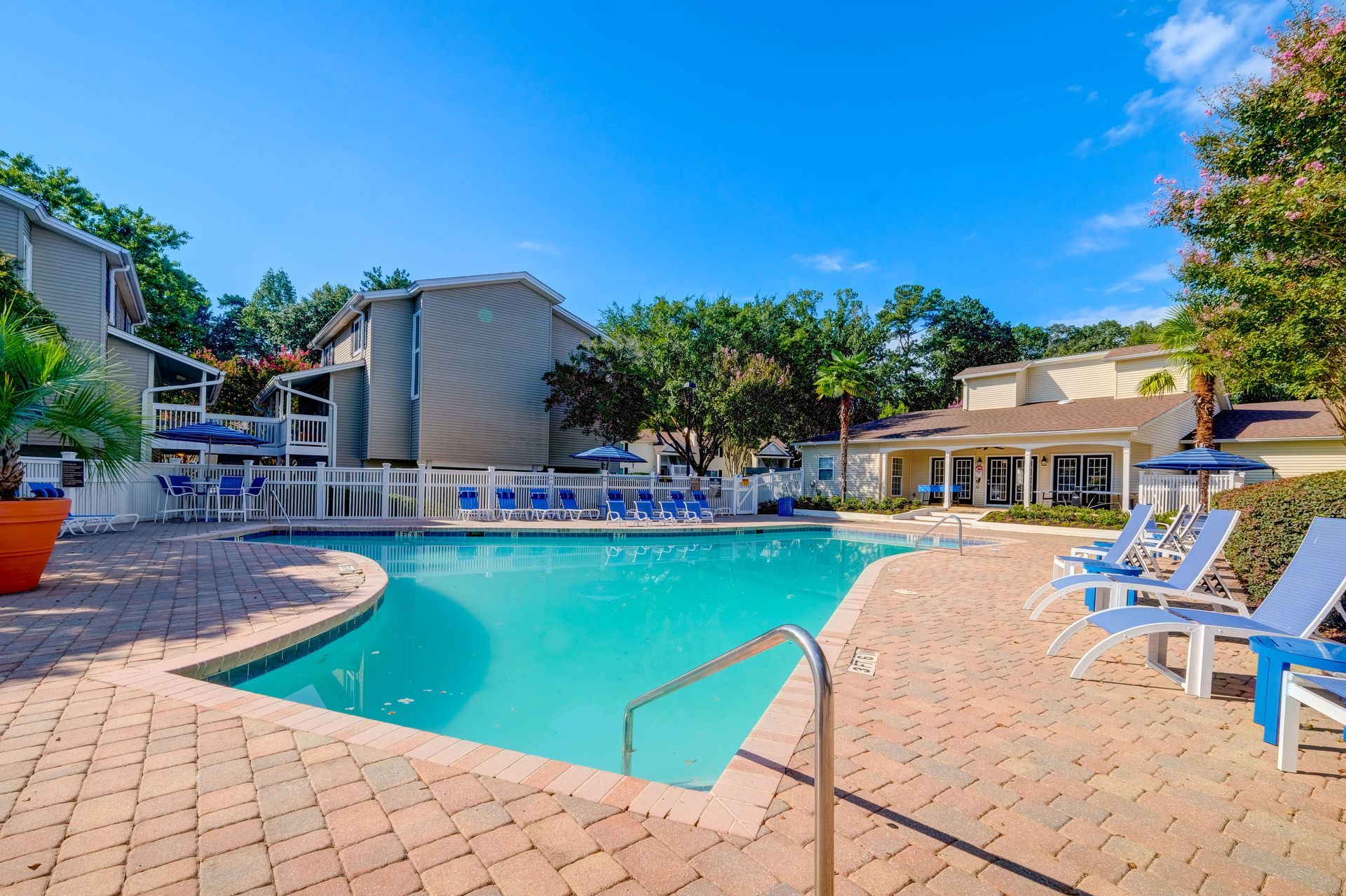 A swimming pool surrounded by brick paving, lounge chairs, and apartment buildings under a clear blue sky.